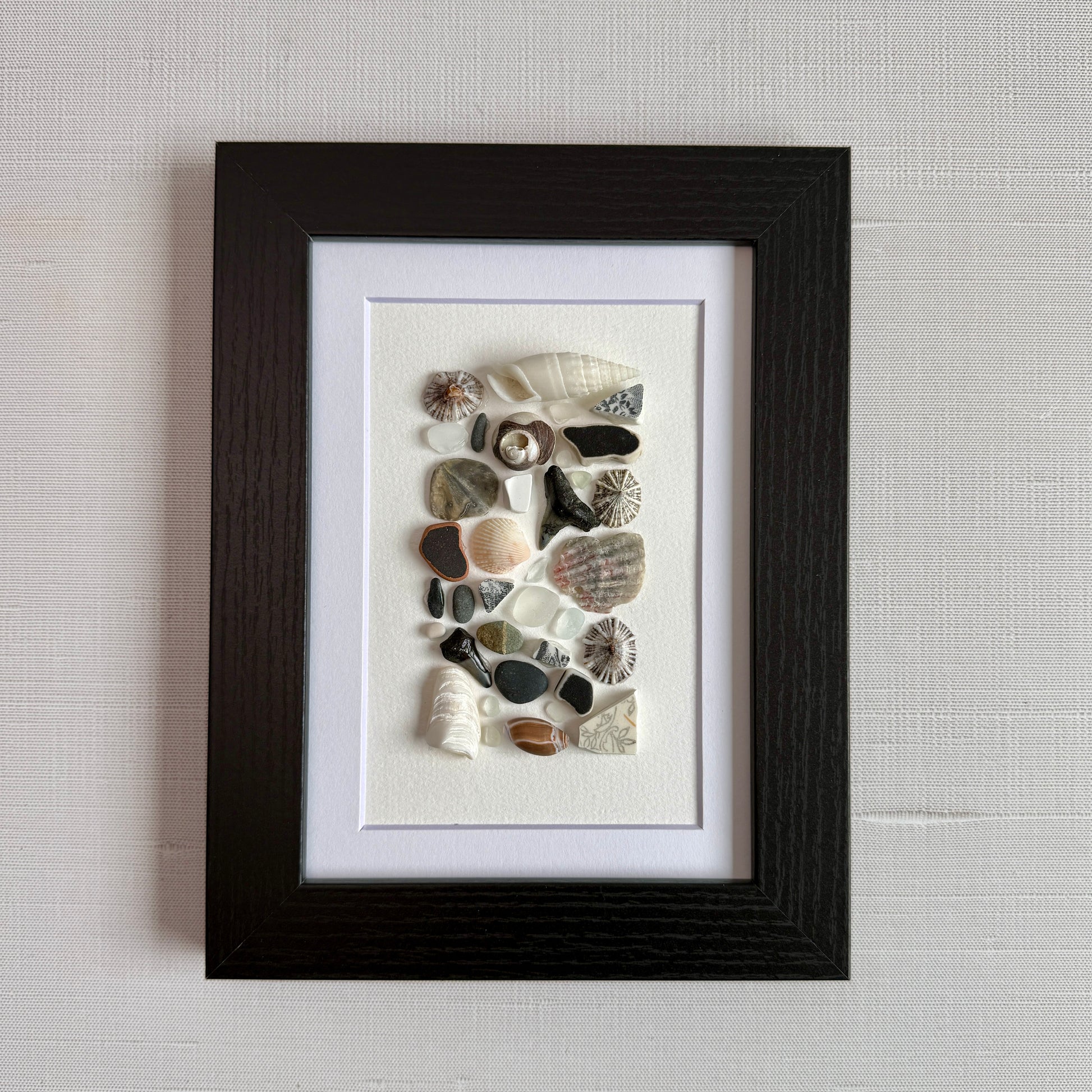 A 5×7 framed wall art piece featuring hand-arranged black, gray and white sea glass and beach-found shells and shark-tooth, in a sleek black wood frame, set against a white background.