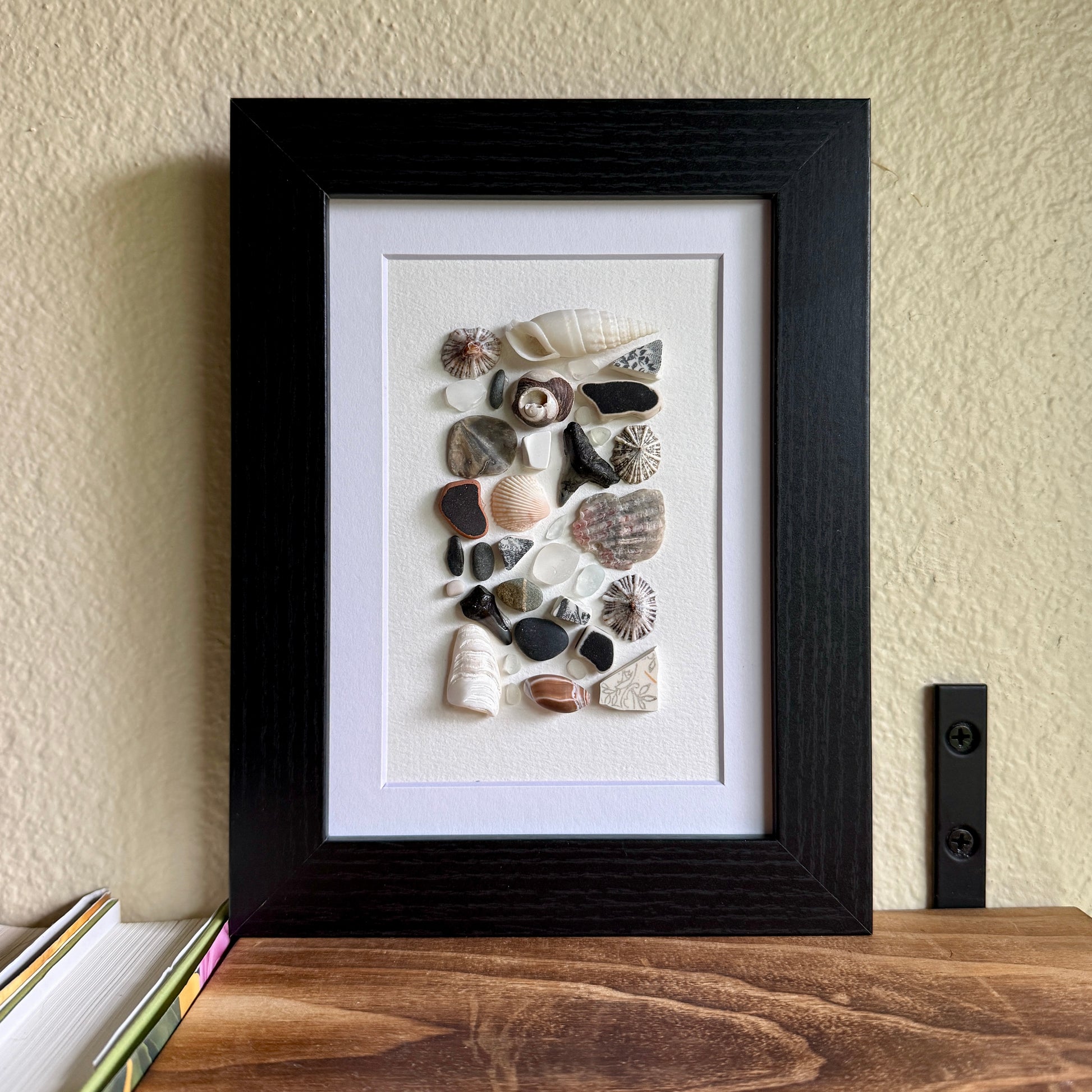 A 5×7 framed wall art piece featuring hand-arranged black, gray and white sea glass and beach-found shells and shark-tooth, in a sleek black wood frame, set against a white background.