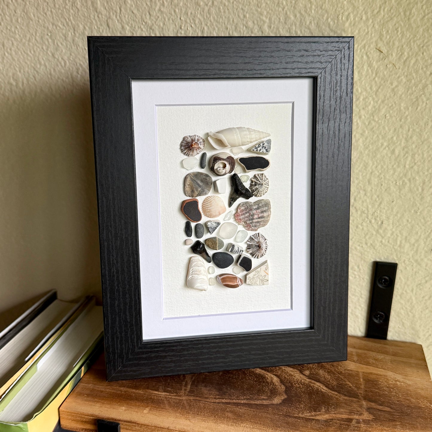 A 5×7 framed wall art piece featuring hand-arranged black, gray and white sea glass and beach-found shells and shark-tooth, in a sleek black wood frame, set against a white background.