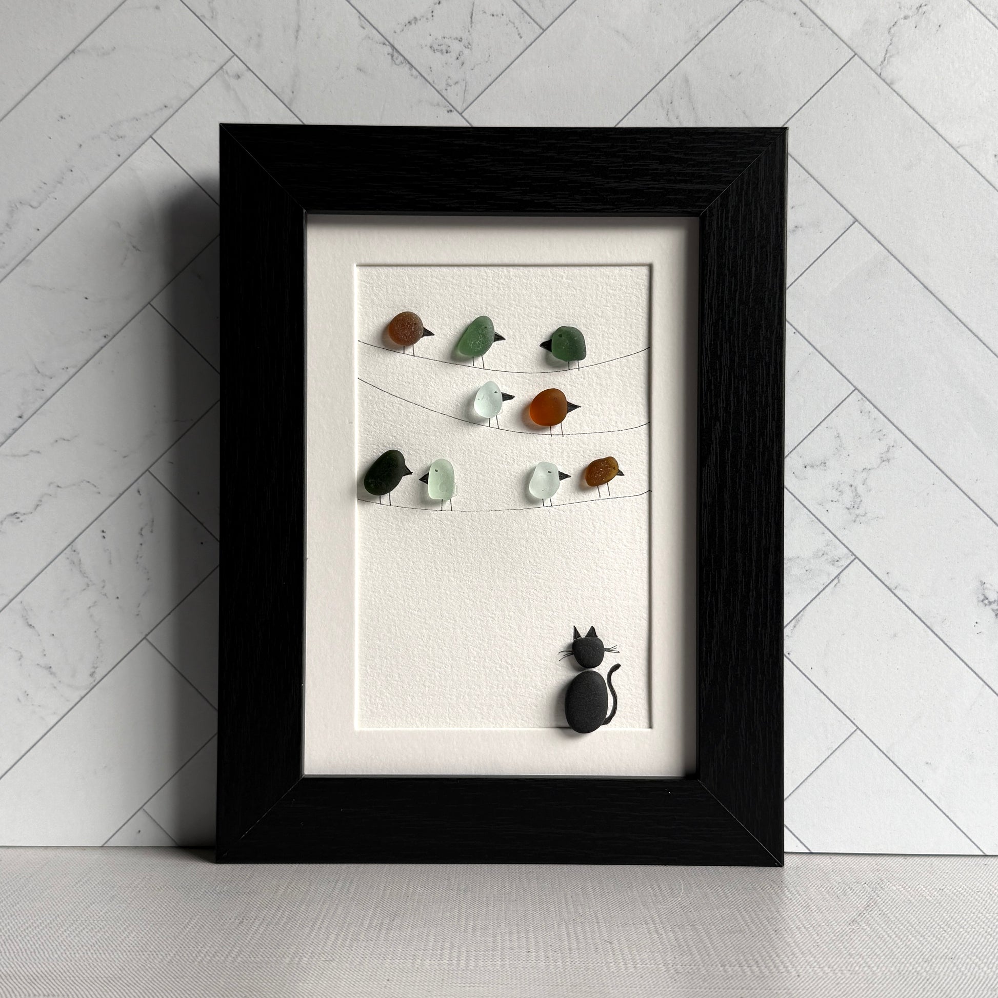 Framed artwork with sea glass birds on telephone wires and a black pebble cat watching from below on a white background