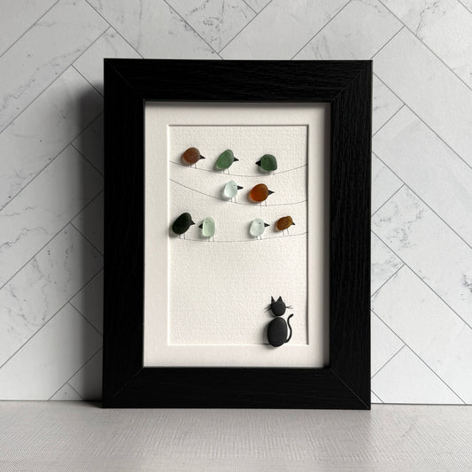 Framed artwork with sea glass birds on telephone wires and a black pebble cat watching from below on a white background