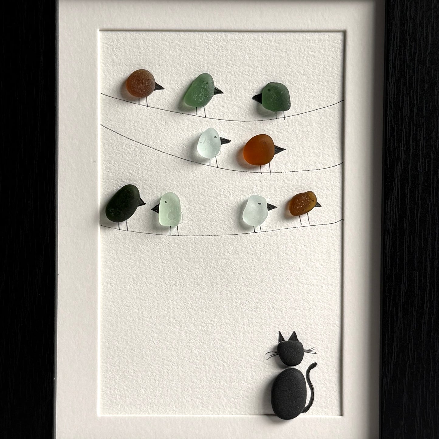Framed artwork with sea glass birds on telephone wires and a black pebble cat watching from below on a white background