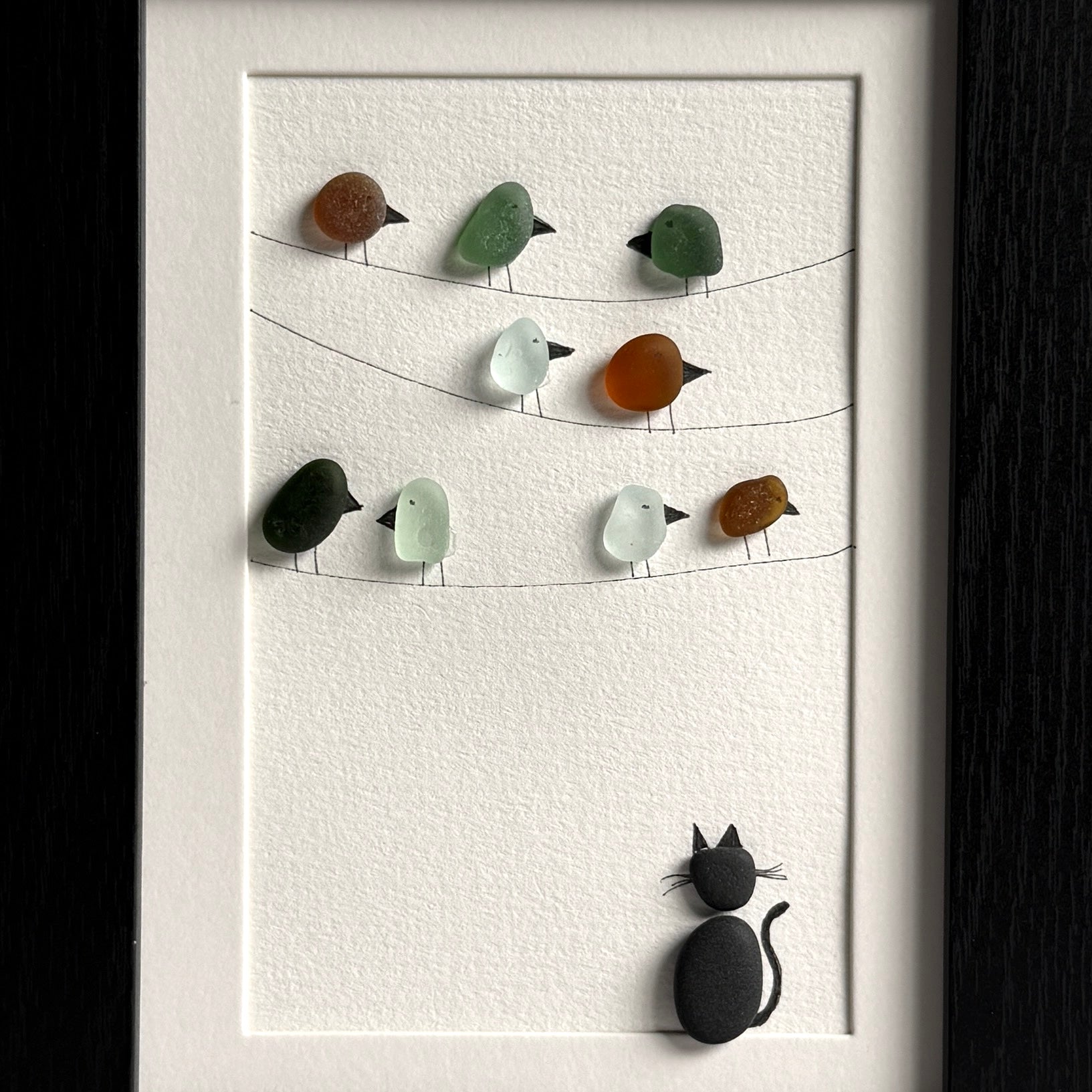 Framed artwork with sea glass birds on telephone wires and a black pebble cat watching from below on a white background