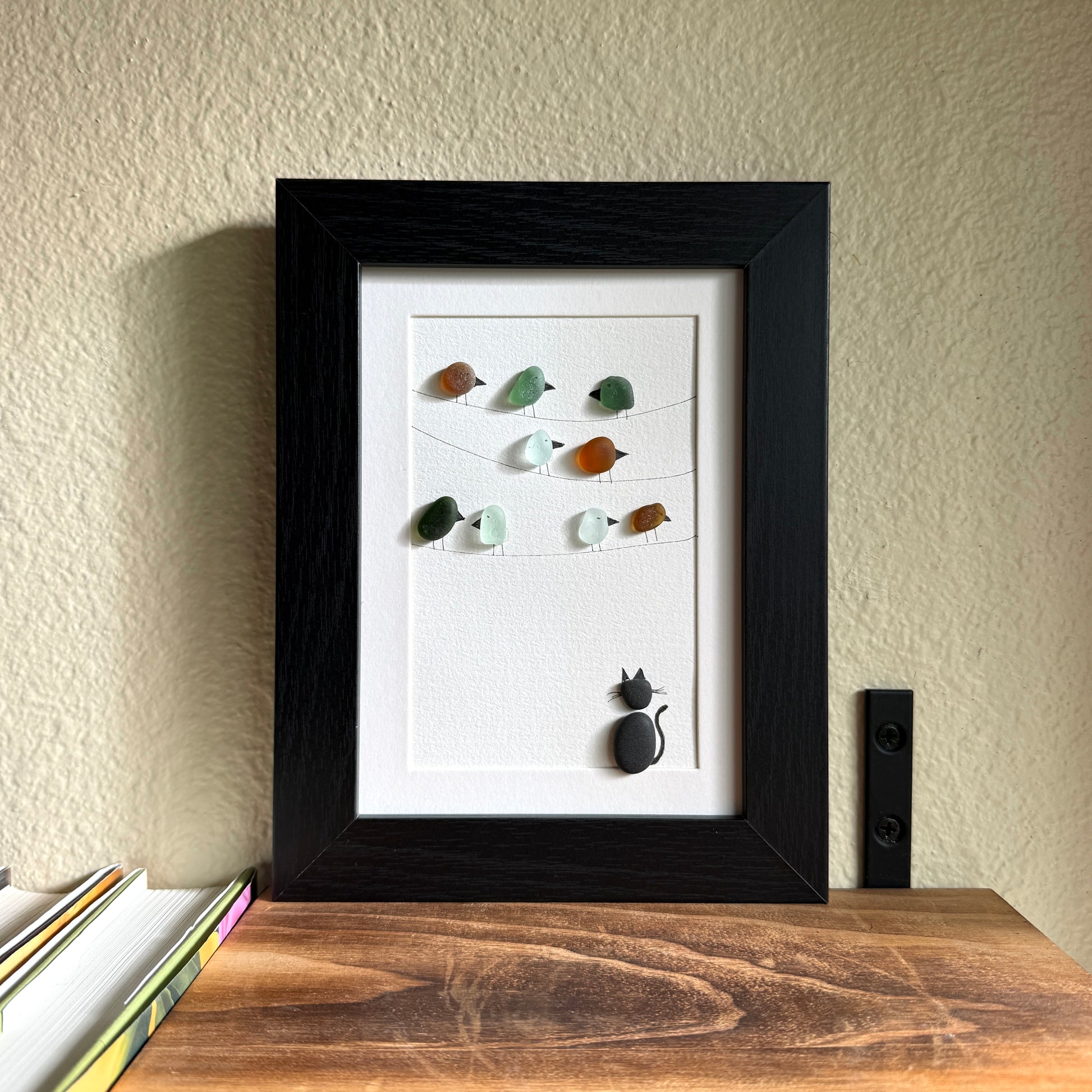 Framed artwork with sea glass birds on telephone wires and a black pebble cat watching from below on a white background
