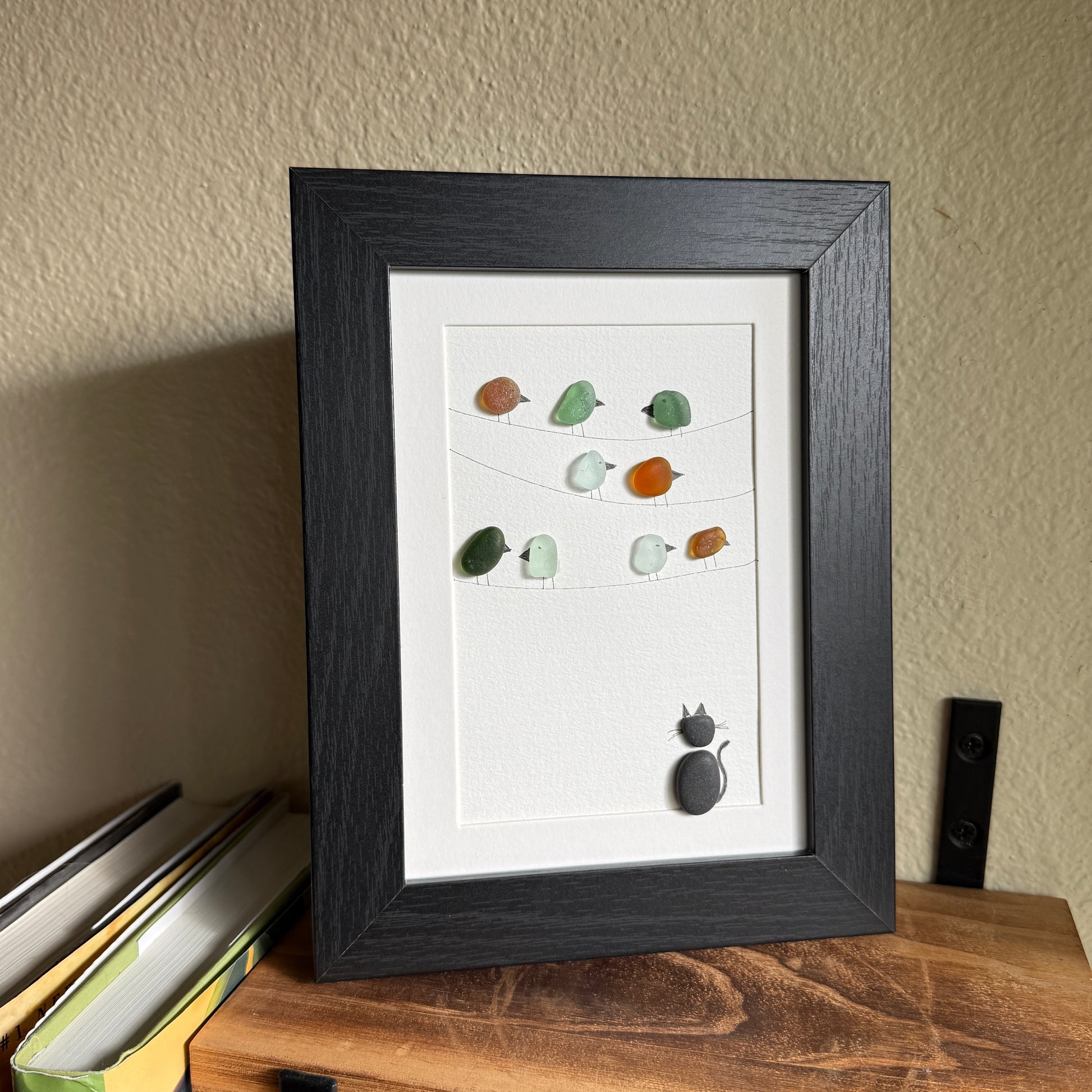 Framed artwork with sea glass birds on telephone wires and a black pebble cat watching from below on a white background