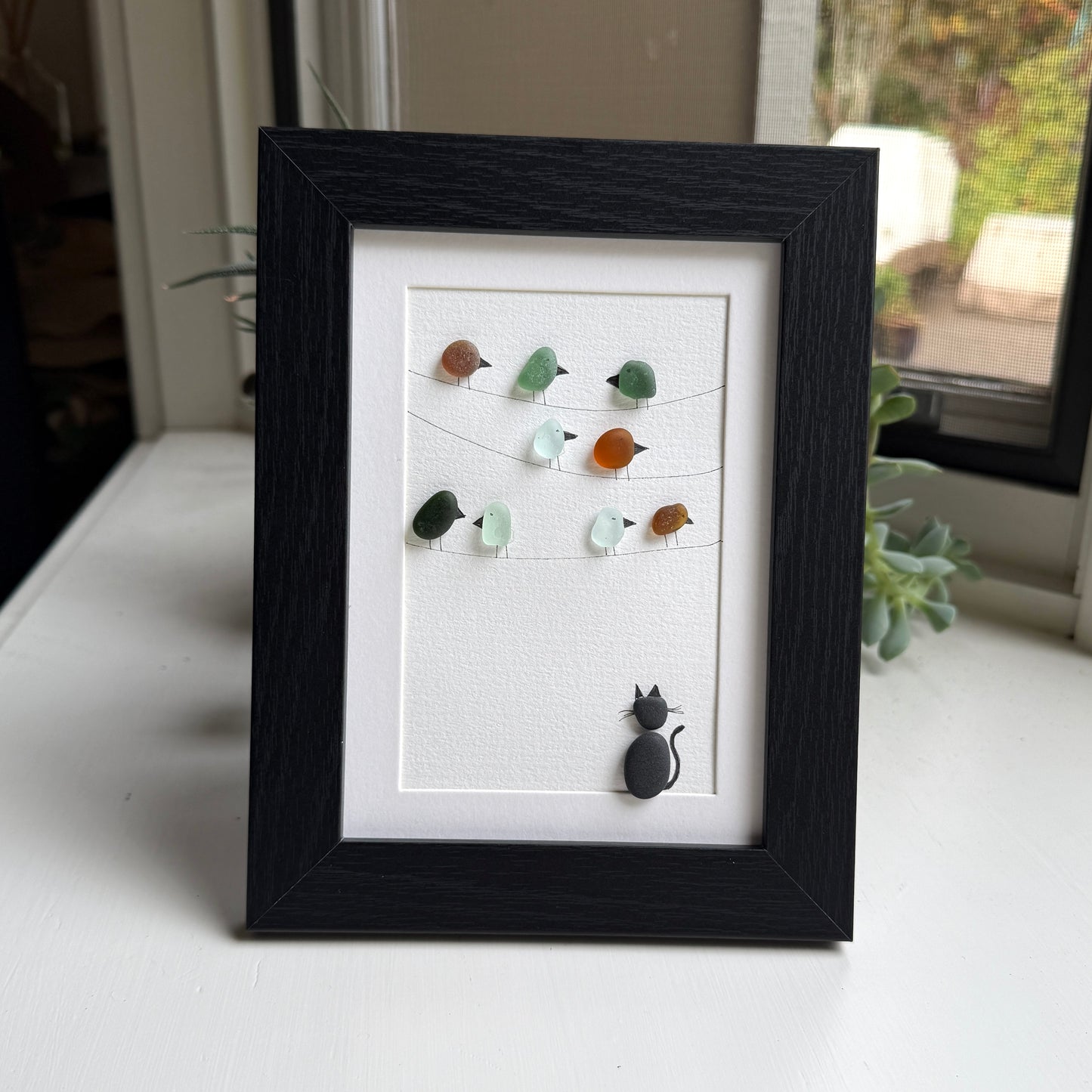 Framed artwork with sea glass birds on telephone wires and a black pebble cat watching from below on a white background