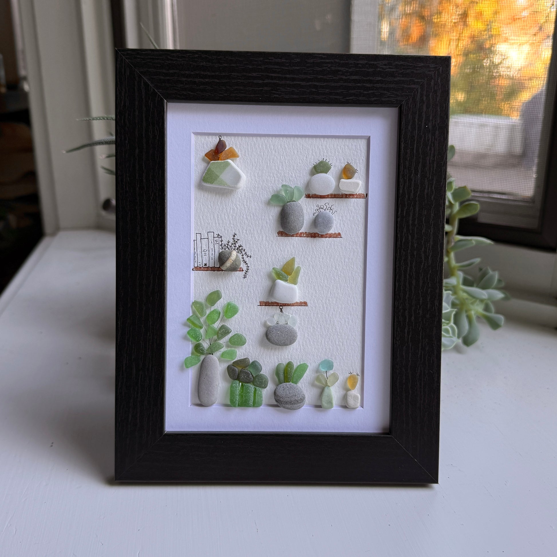 Framed 5x7 sea glass and pebble  artwork of small plants and a cat on shelves against a white background