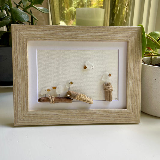 This is a framed 5 x 7 inch sea glass art picture featuring seagulls and driftwood logs. This item is perfect for gift giving and is ready to hang or place on a shelf. 