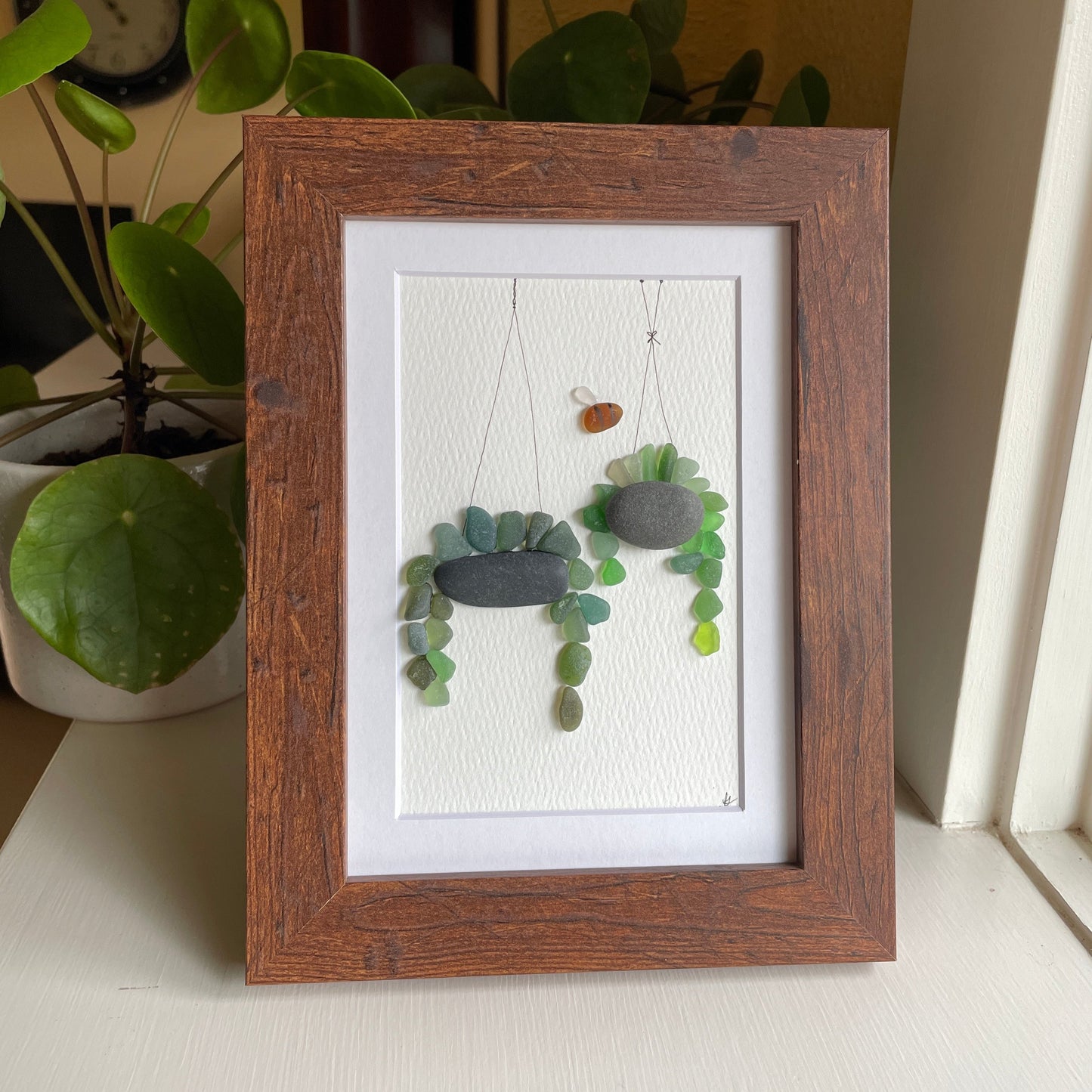 This 5 x 7 inch framed sea glass art picture features two hanging plants made from genuine sea glass, pottery & pebbles with a cute little sea glass bee buzzing about.  Designs may vary. 