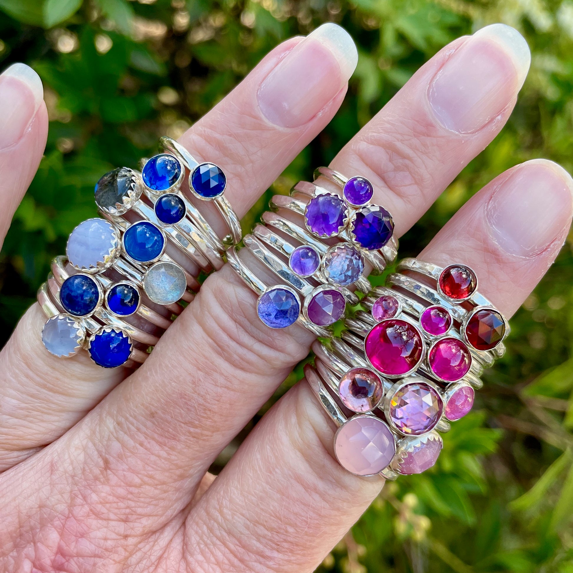 Hand wearing multiple rings with colorful gemstones against a blurred green background