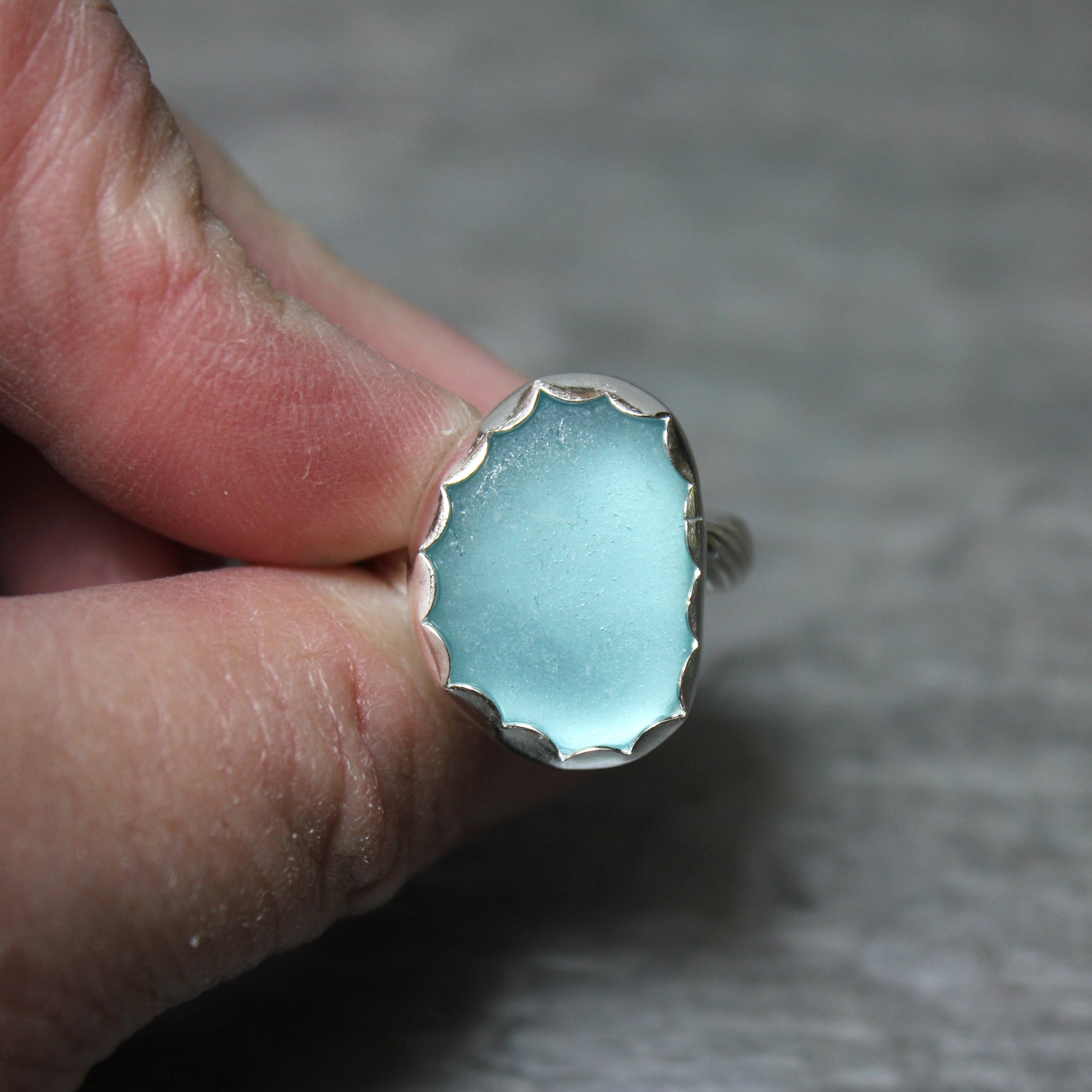 Hand holding a ring with an aqua sea glass and silver band against a neutral background