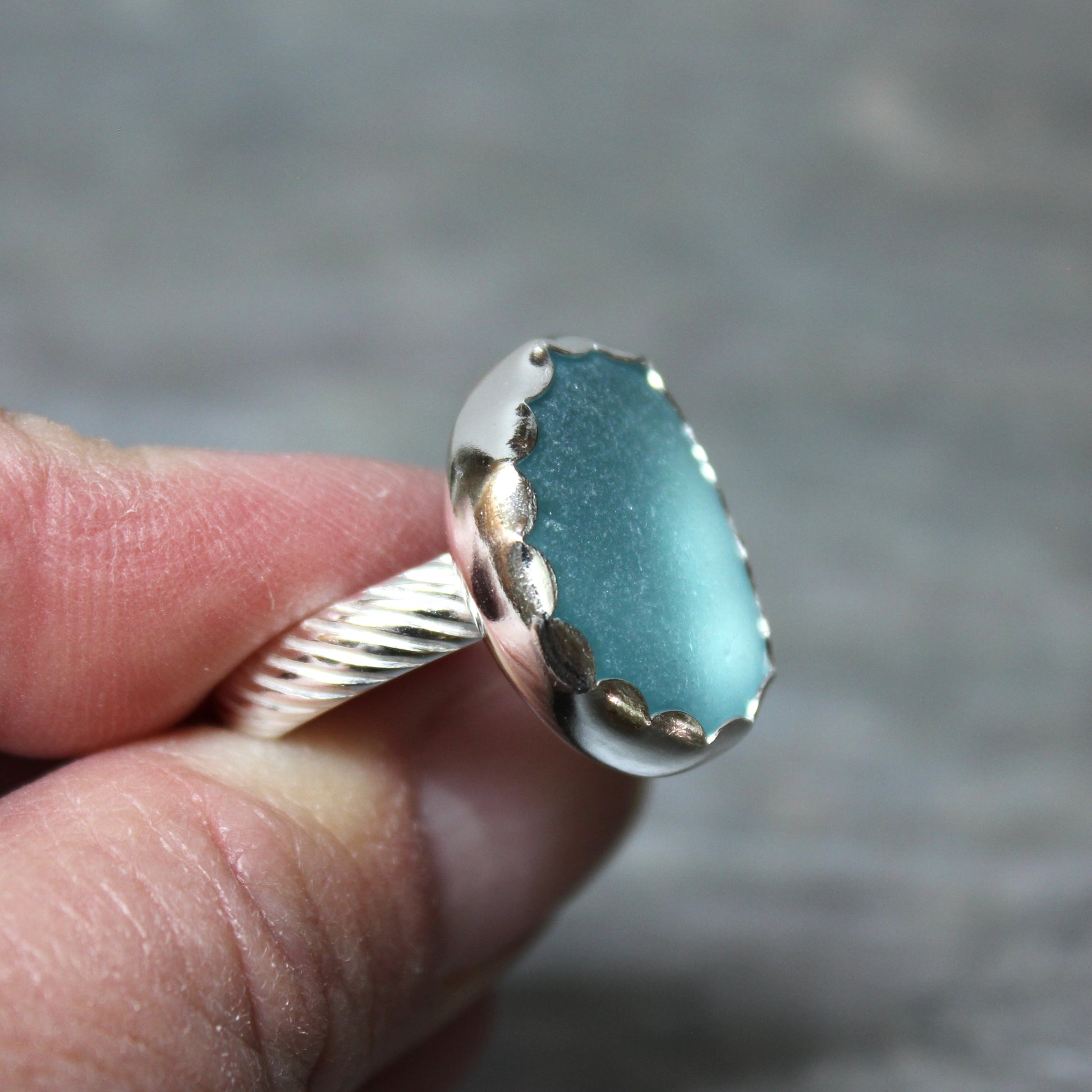 Aqua Sea Glass ring with silver band held between fingers against a blurred background