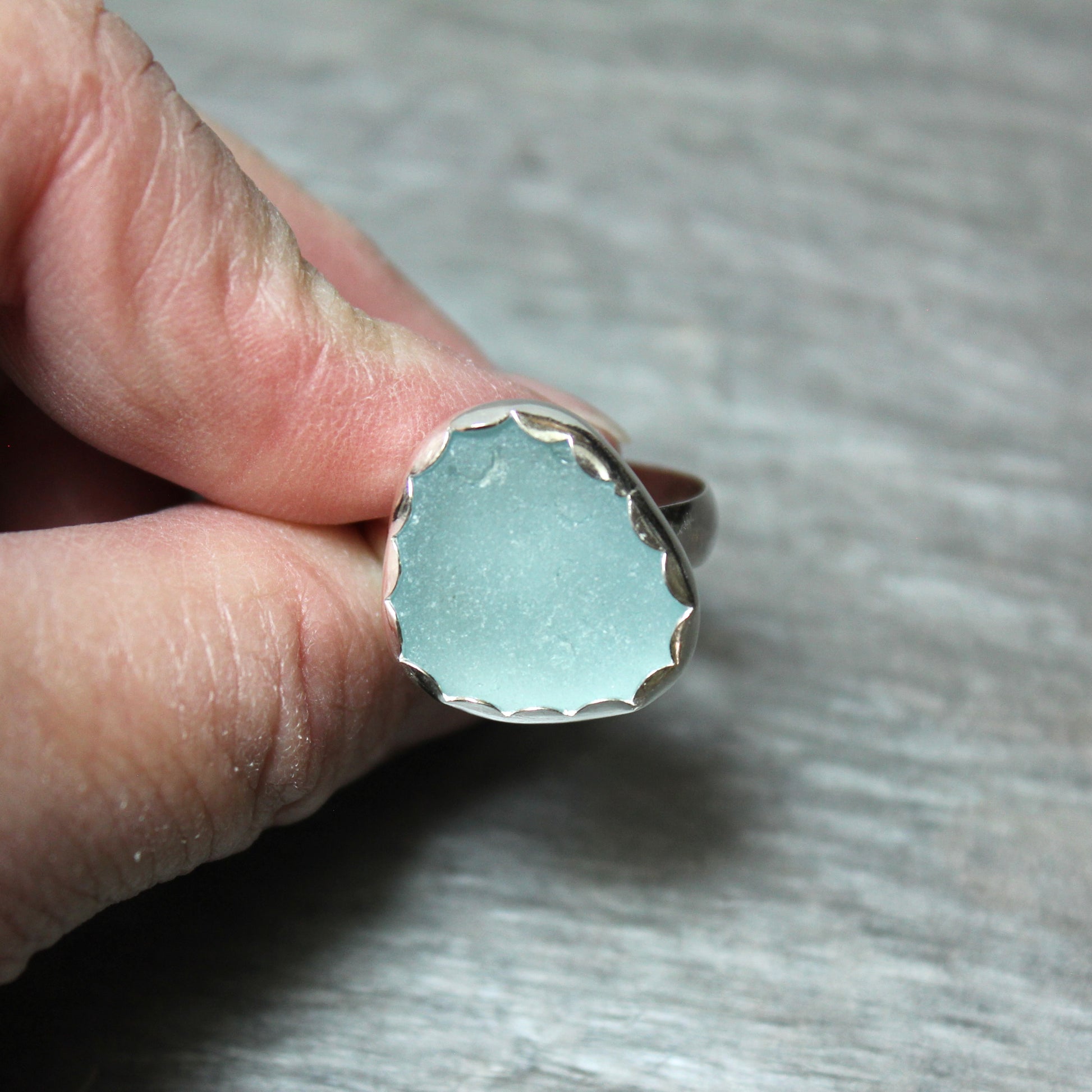 Hand holding a ring with a light blue sea glass and silver band on a neutral background