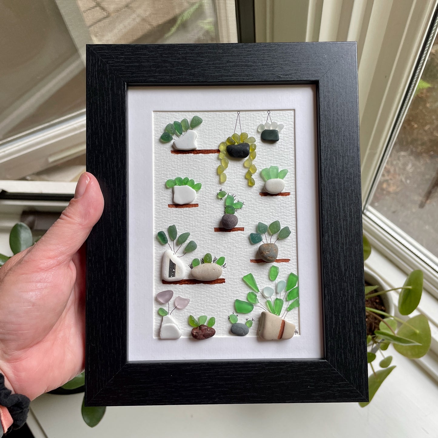 This is a framed 5 x 7 inch sea glass art picture featuring several potted succulents, cactus and plants made with sea glass and beach pottery. This item is perfect for gift giving and ready to hang or place on a shelf.  