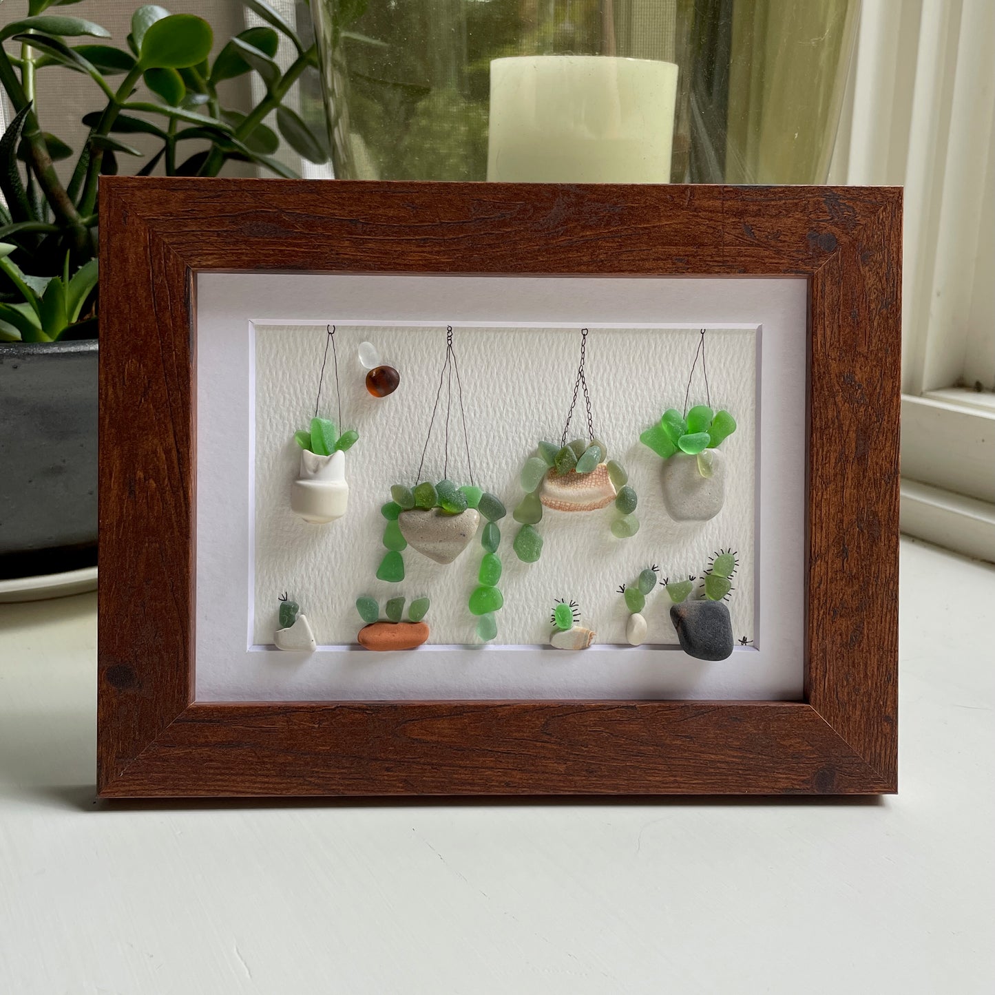 This is a framed 5 x 7 inch sea glass art picture featuring several potted succulents, cactus and plants made with sea glass and beach pottery. This item is perfect for gift giving and ready to hang or place on a shelf.  