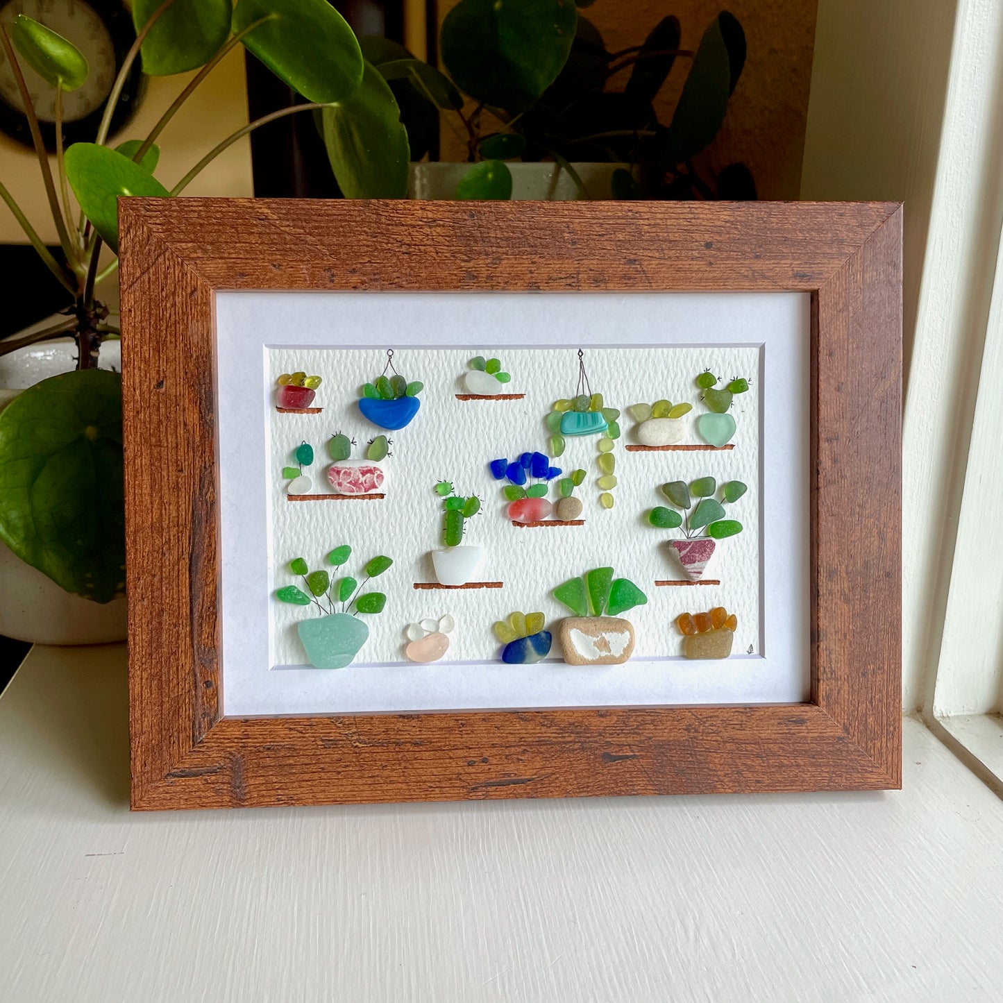 This is a framed 5 x 7 inch sea glass art picture featuring several potted succulents, cactus and plants made with sea glass and beach pottery. This item is perfect for gift giving and ready to hang or place on a shelf.  