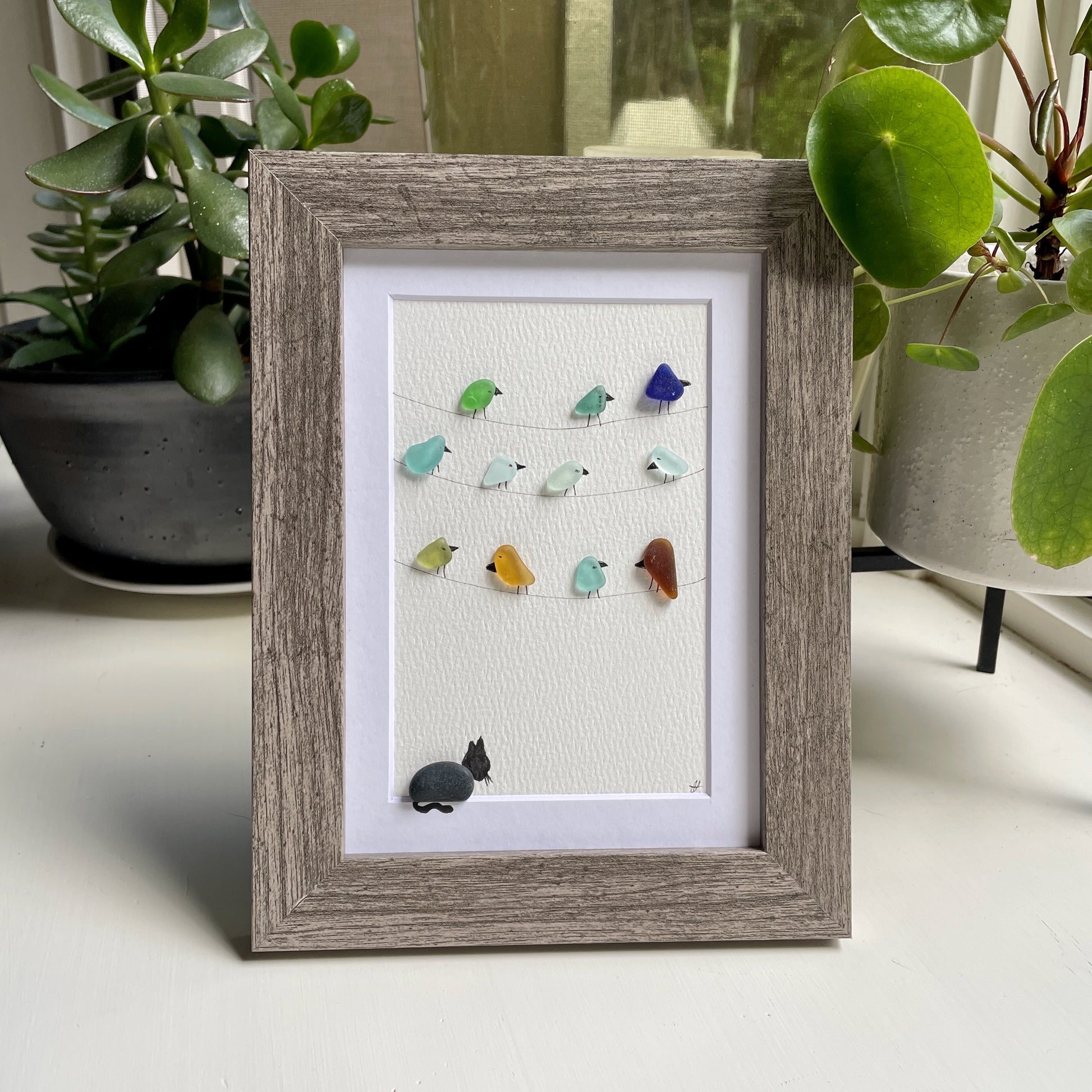 This is a framed 5 x 7 inch sea glass art picture featuring several birds perched on wires and a pebble cat sitting below watching. This item makes a perfect gift for a cat lover or bird lover and is ready to hang or place on a shelf. 