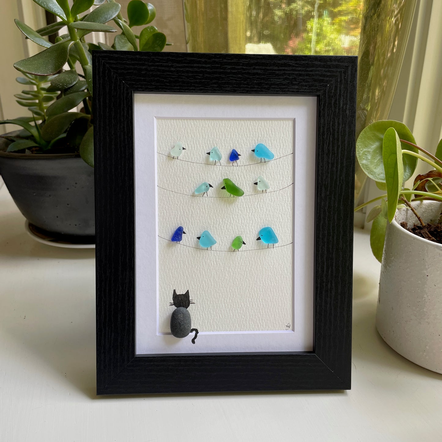 This is a framed 5 x 7 inch sea glass art picture featuring several birds perched on wires and a pebble cat sitting below watching. This item makes a perfect gift for a cat lover or bird lover and is ready to hang or place on a shelf. 