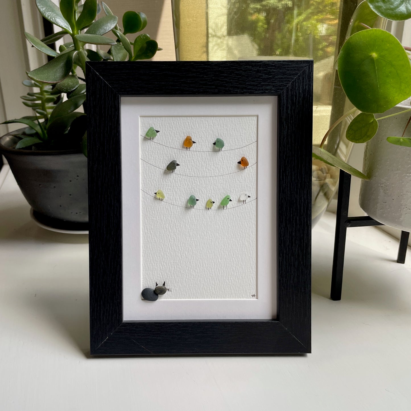 This is a framed 5 x 7 inch sea glass art picture featuring several birds perched on wires and a pebble cat sitting below watching. This item makes a perfect gift for a cat lover or bird lover and is ready to hang or place on a shelf. 