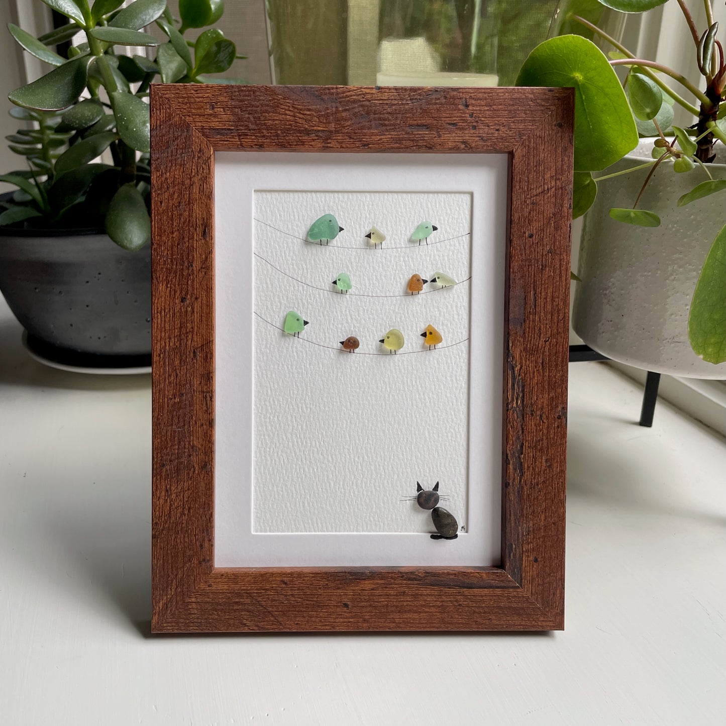 This is a framed 5 x 7 inch sea glass art picture featuring several birds perched on wires and a pebble cat sitting below watching. This item makes a perfect gift for a cat lover or bird lover and is ready to hang or place on a shelf. 