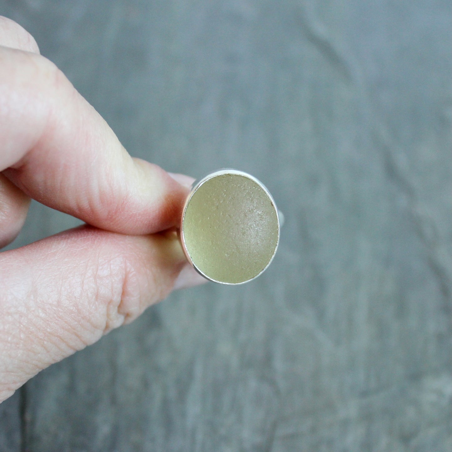 Hand holding a silver ring with a Champagne Sea Glass setting on a gray background