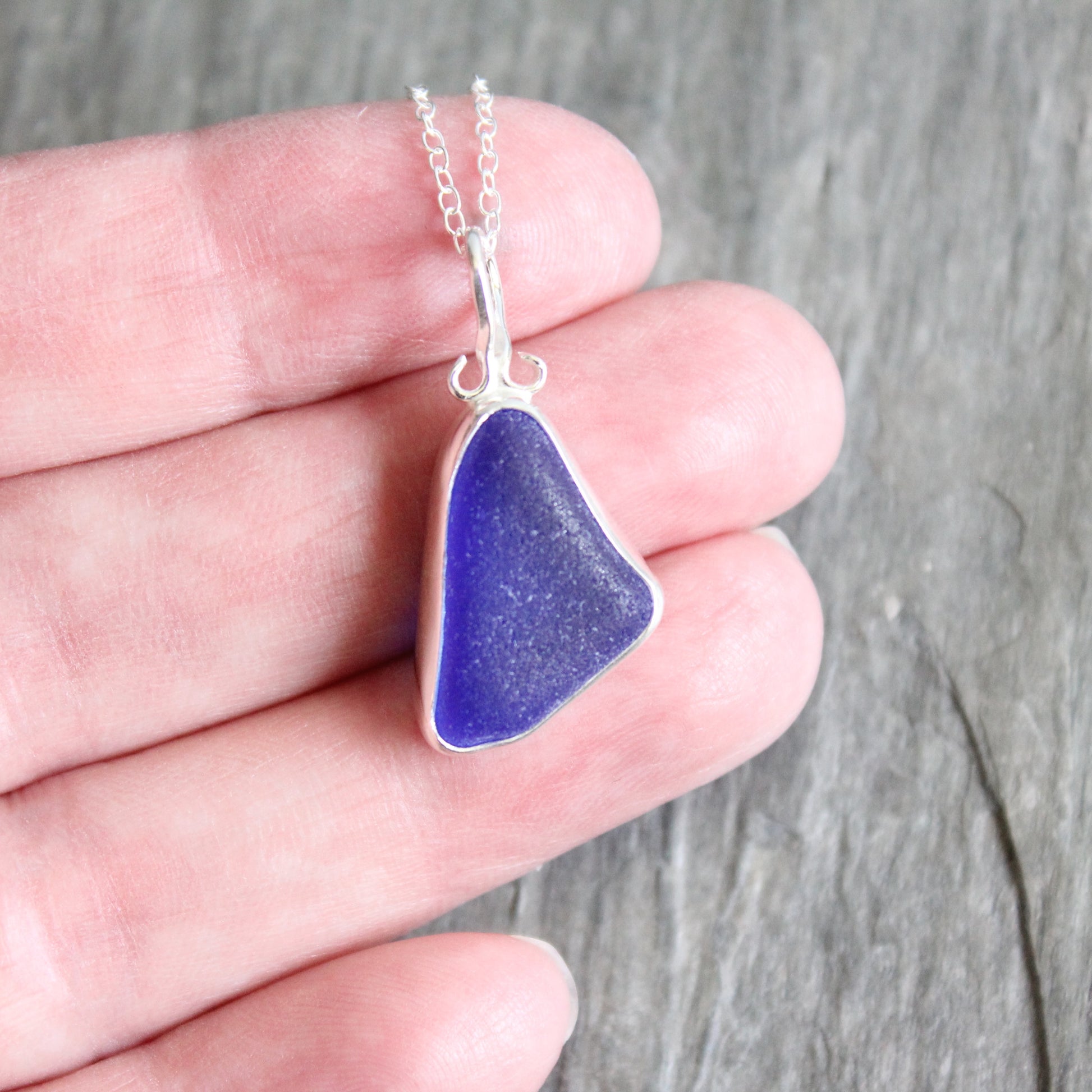 Cobalt sea glass pendant on a silver chain held in a hand against a wooden background
