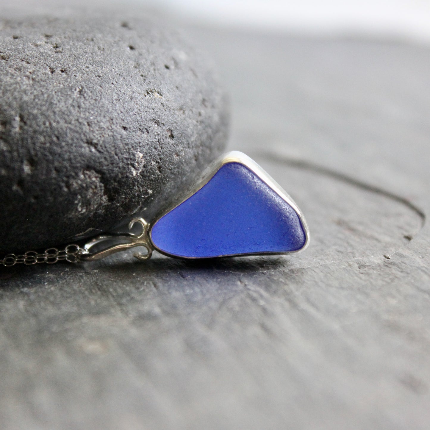 Cobalt blue sea glass triangular pendant on a chain against a gray stone background