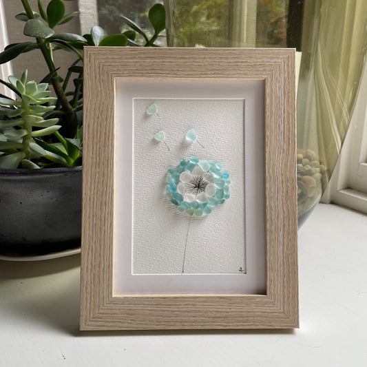 this is a framed 5 x 7 inch sea glass art picture featuring a dandelion made from aqua and white sea glass. This item is framed and perfect for gift giving and is ready to hang on a wall. 