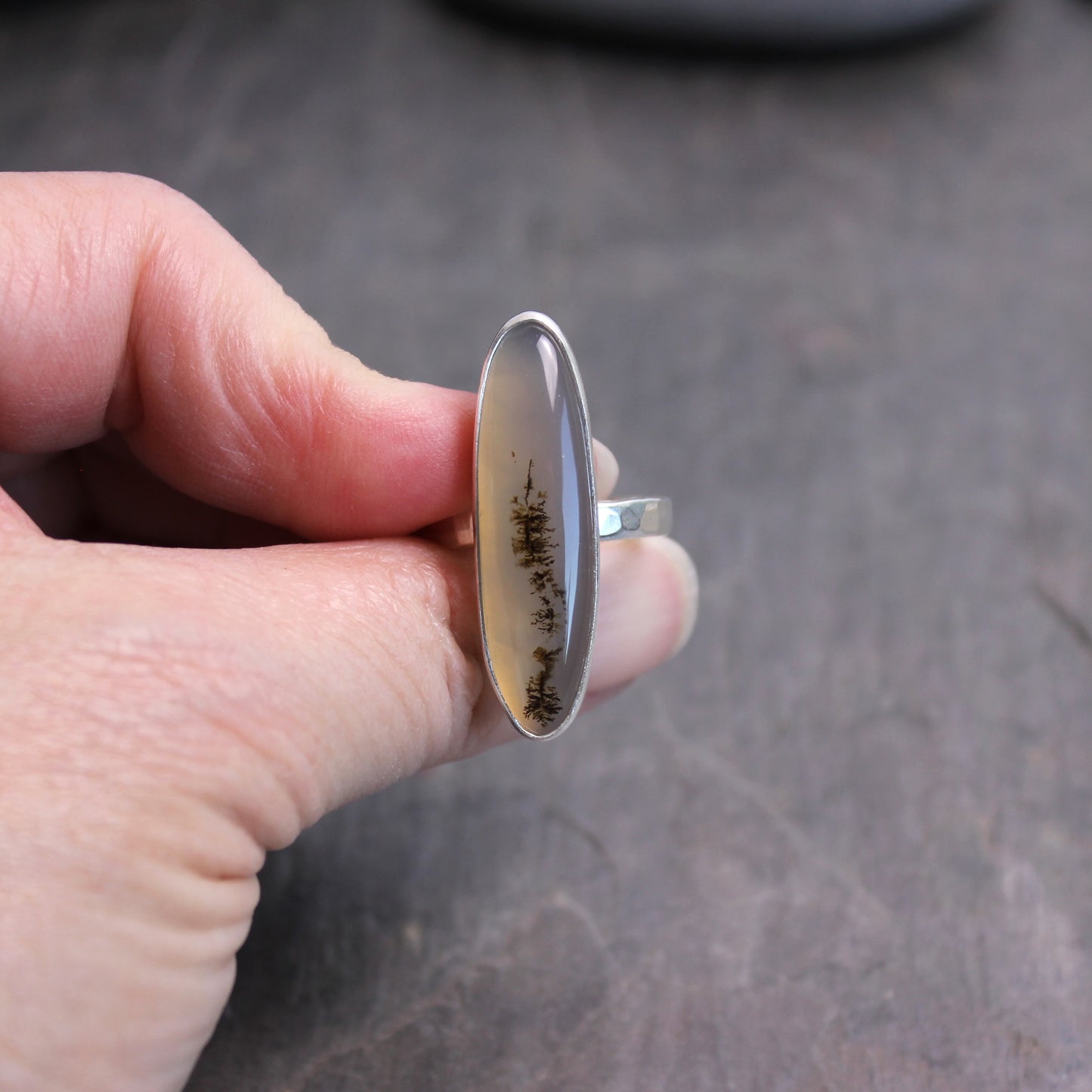 Hand holding a ring with an oval stone featuring tree patterns on a dark surface