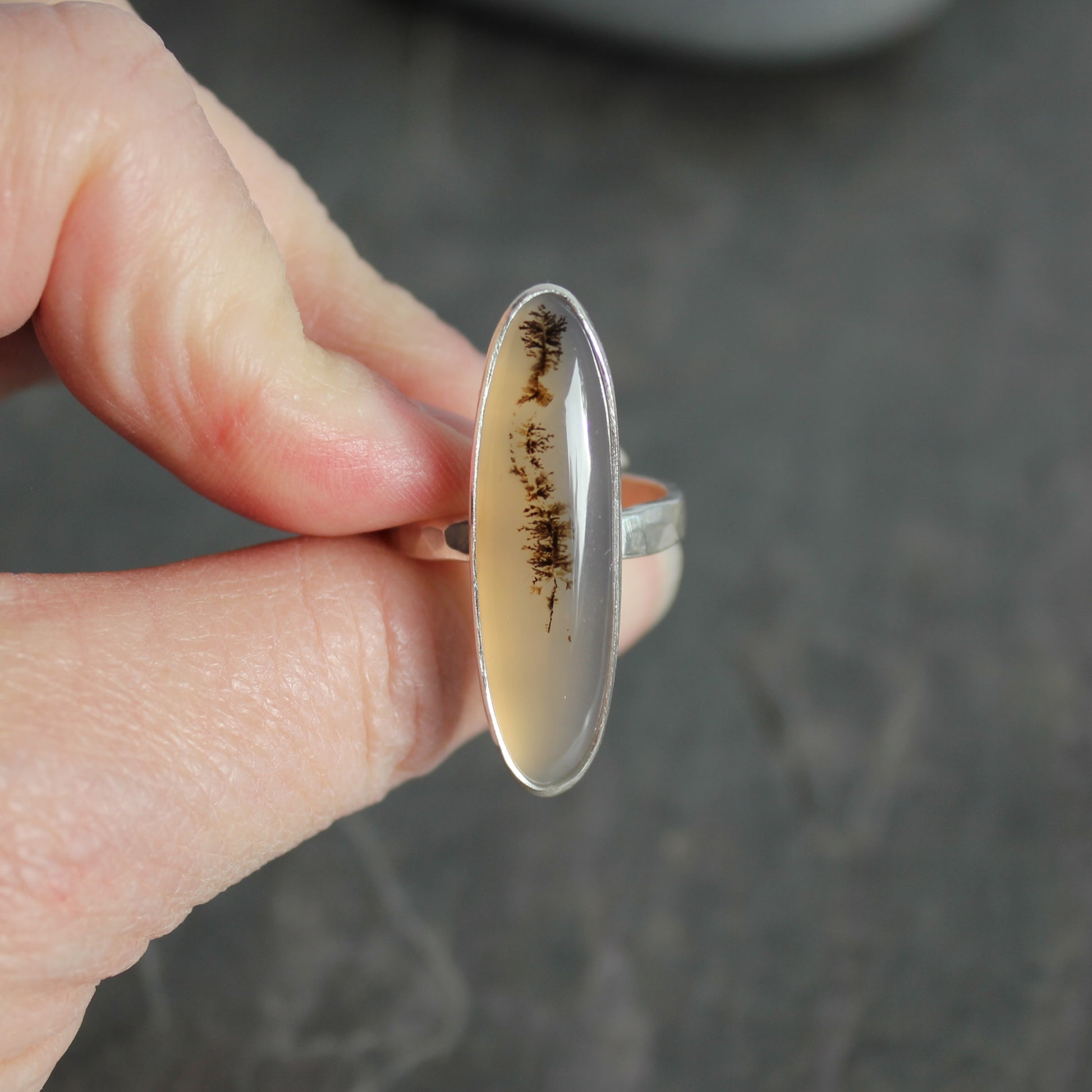 Silver ring with an oval stone held by a hand against a dark background