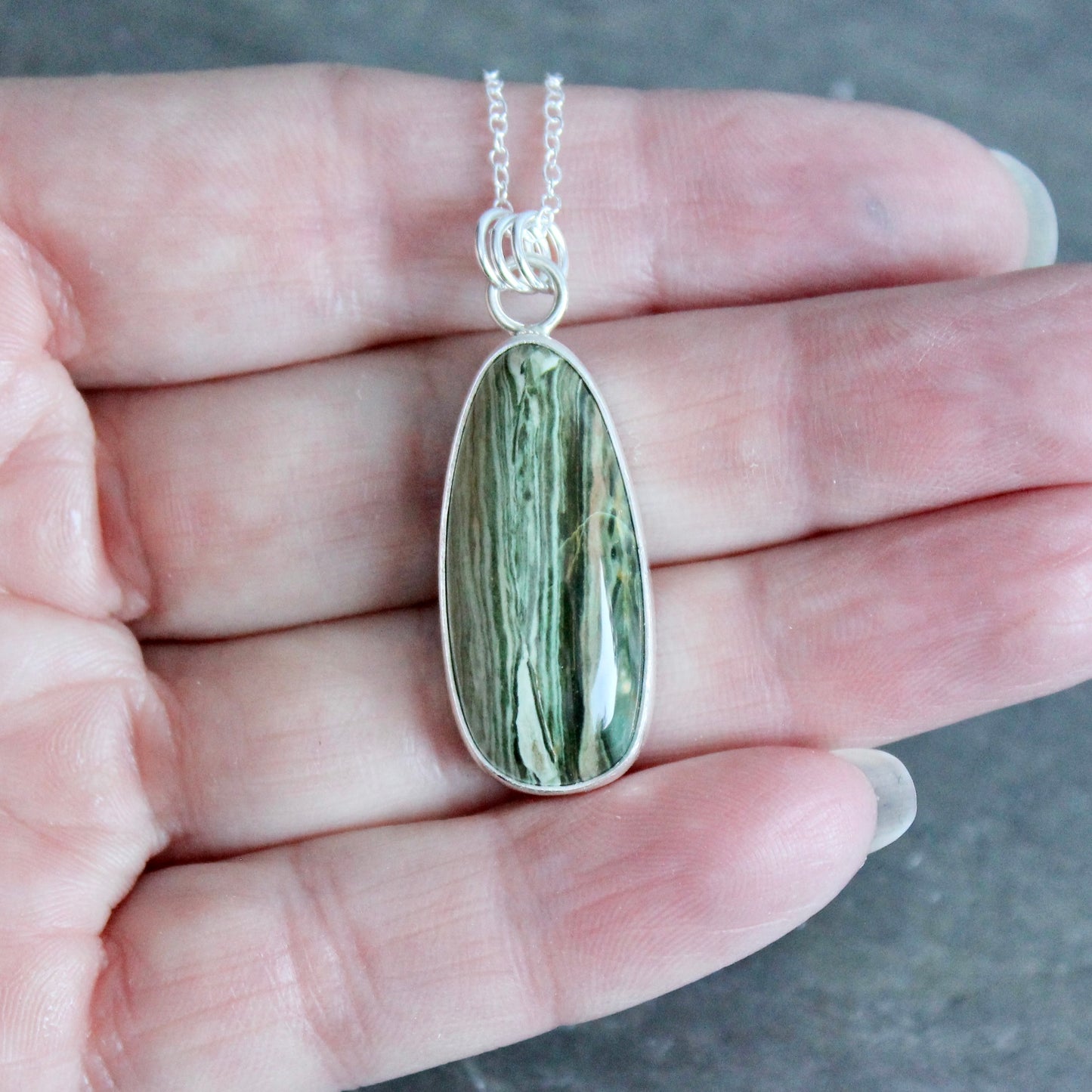 Teardrop-shaped green stone pendant on a silver chain held in a hand.