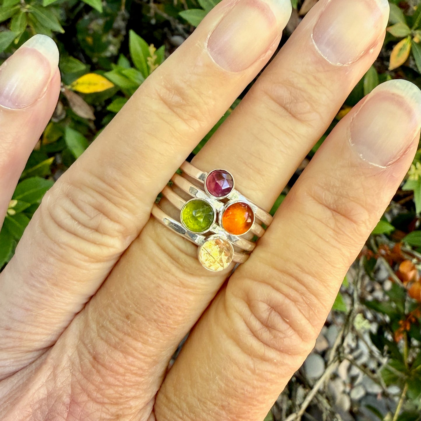 Close-up of a hand wearing a silver ring with colorful gemstones against a natural background.
