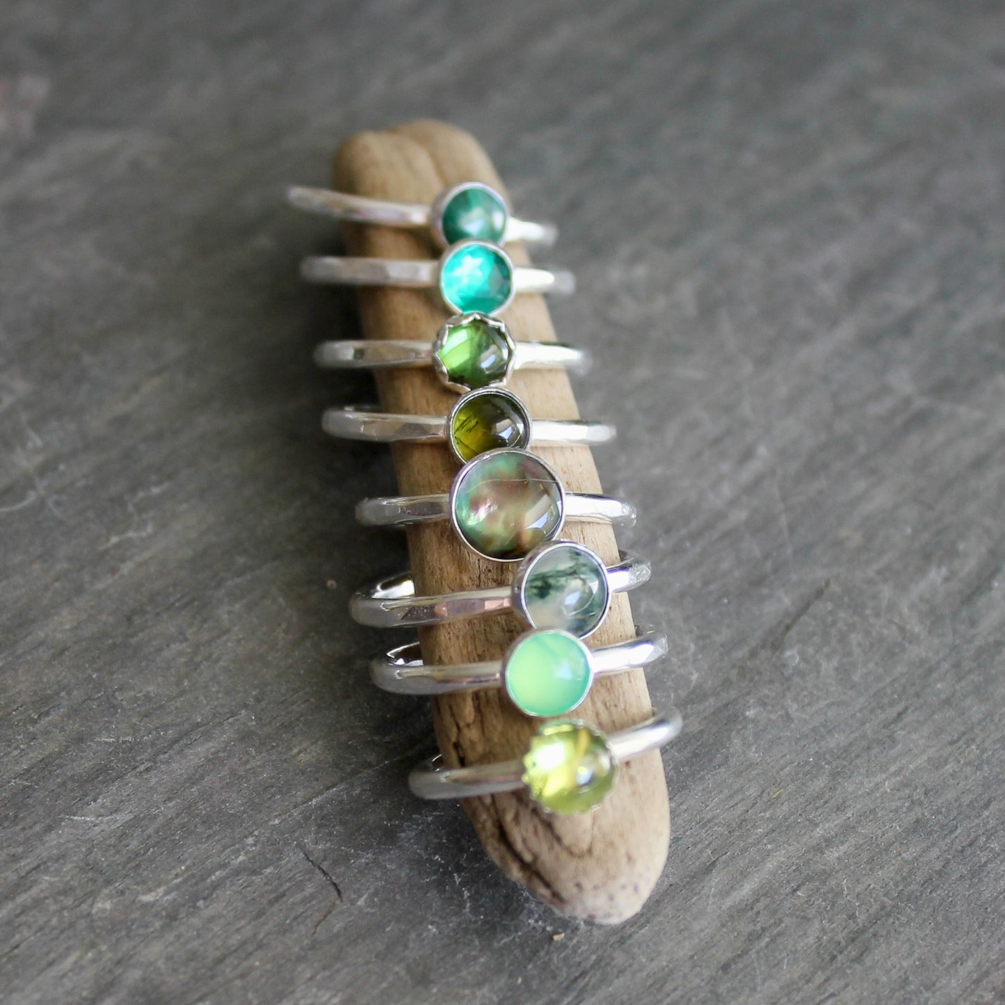 Stack of silver rings with green gemstones on a piece of driftwood against a gray background