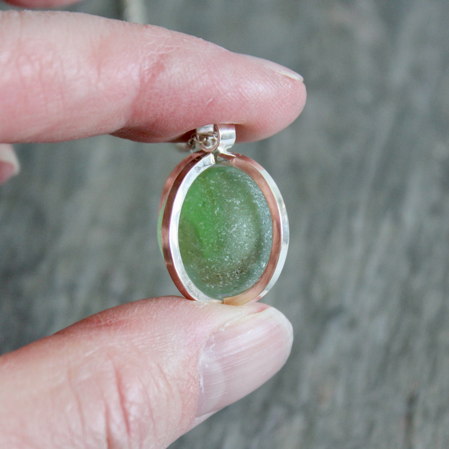 Green Japanese Codd Bottle Sea glass pendant with silver frame held between fingers against a blurred background