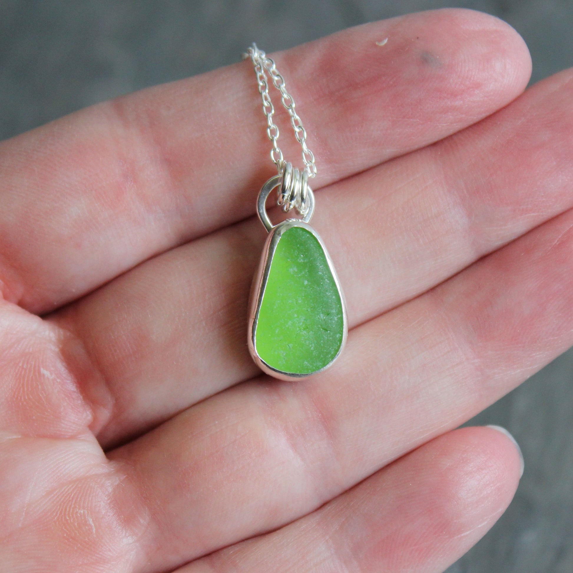Green sea glass teardrop pendant on a silver chain held in a hand.