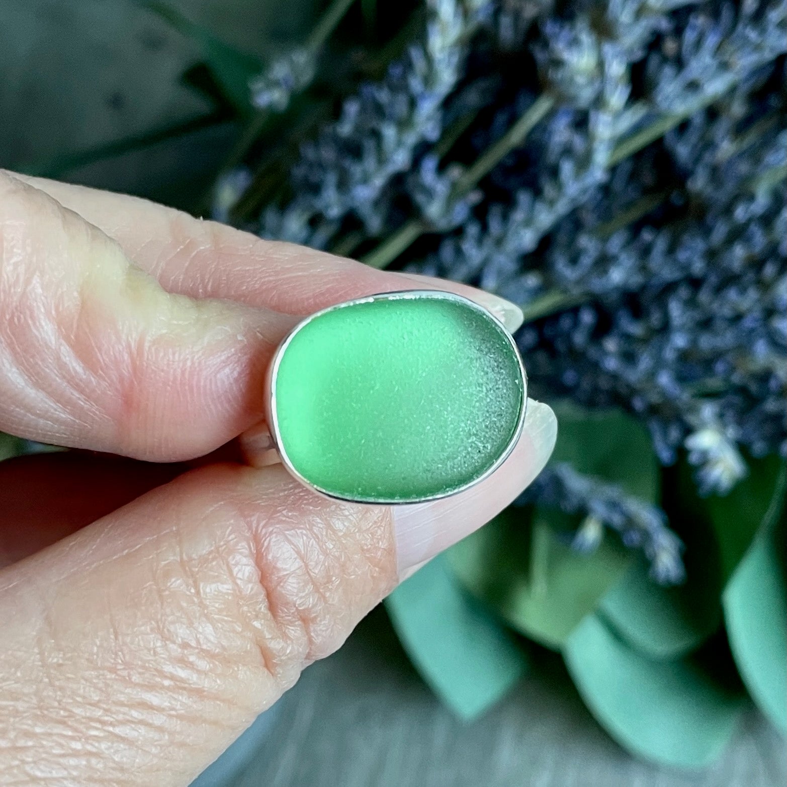 Green sea glass ring held between fingers with a blurred natural background