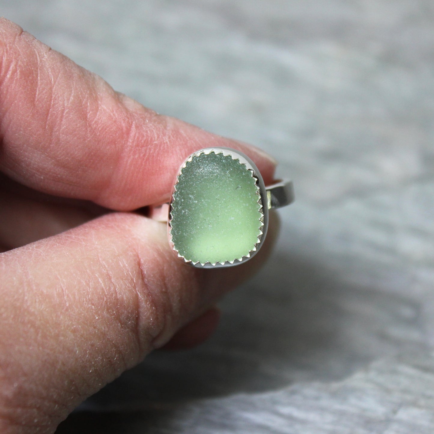 Green sea glass ring with silver setting held on a finger against a neutral background
