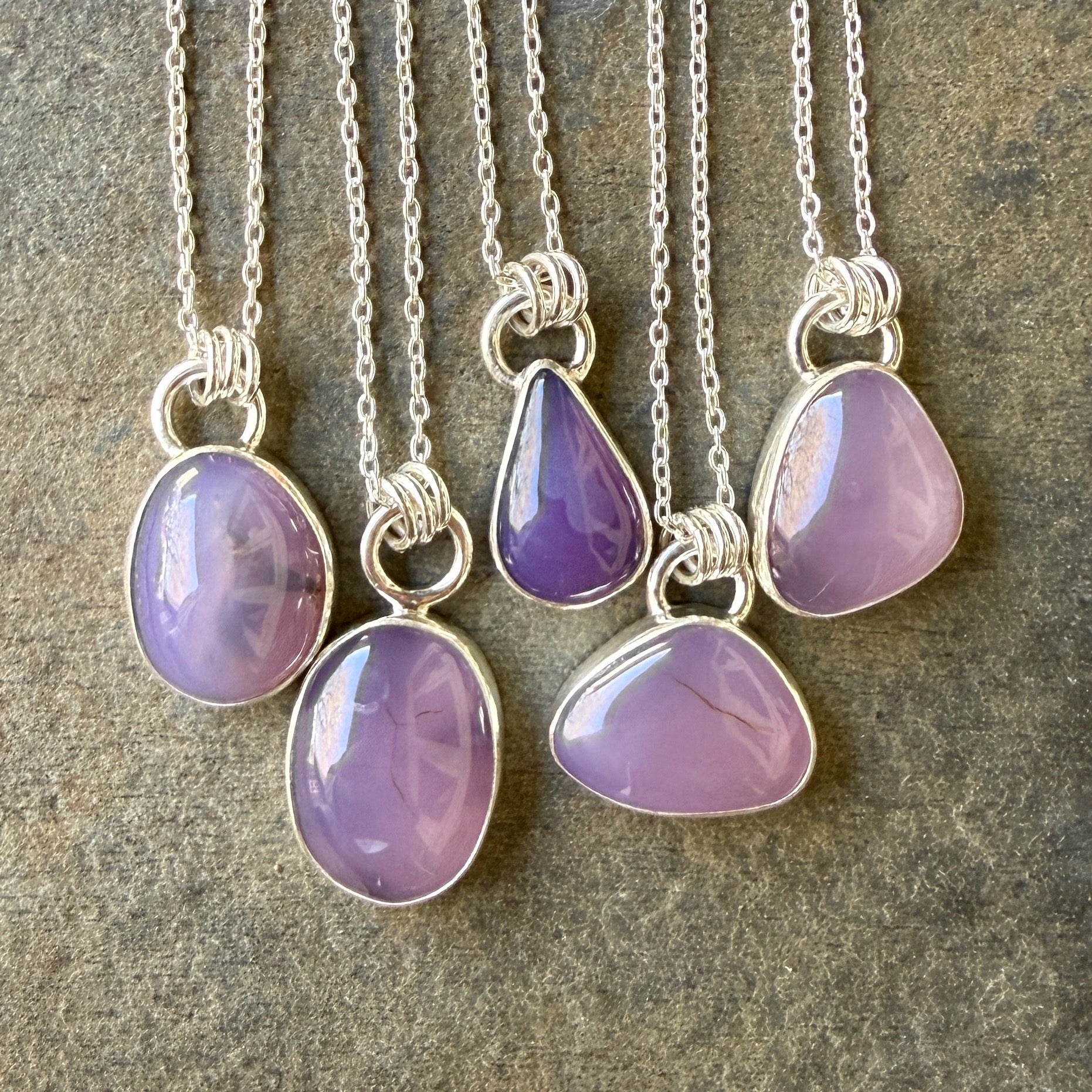 Five silver necklaces with purple teardrop pendants on a textured surface