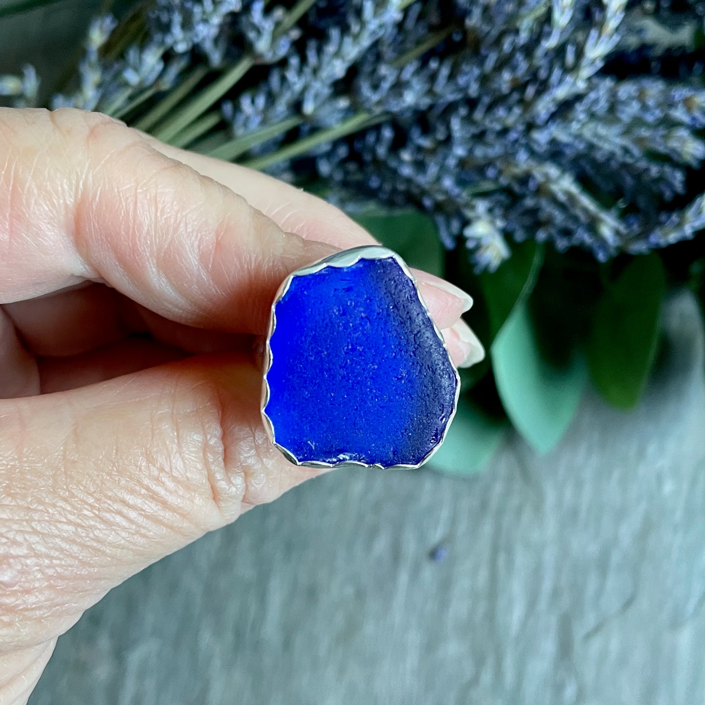 Hand holding a blue sea glass ring with a background of greenery