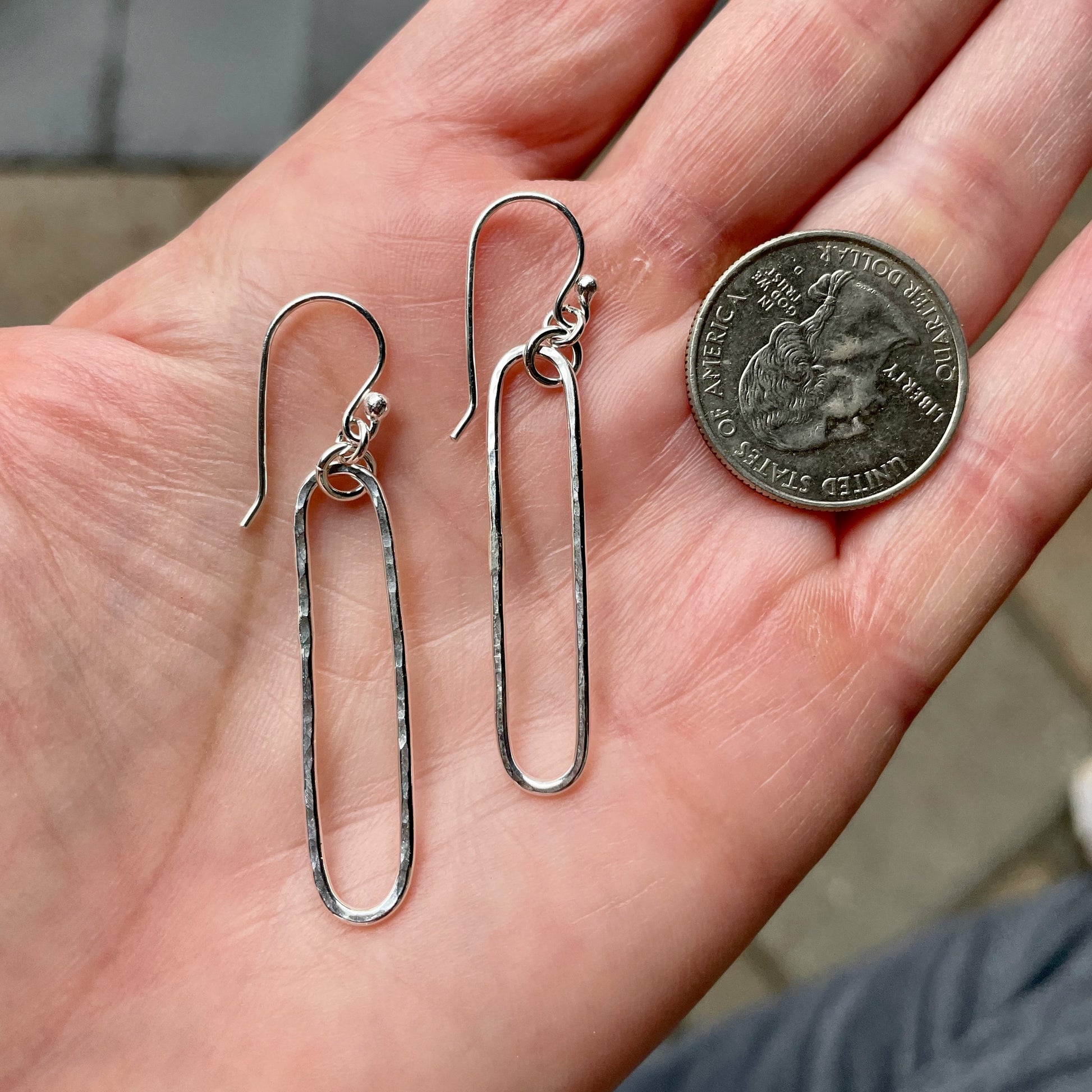 Silver earrings on a hand with a quarter for scale.