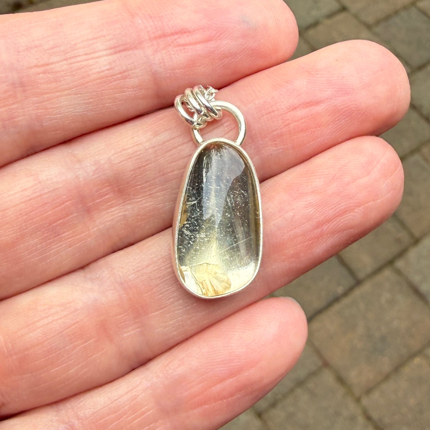 Silver pendant with a clear stone held in a hand on a blurred pavement background