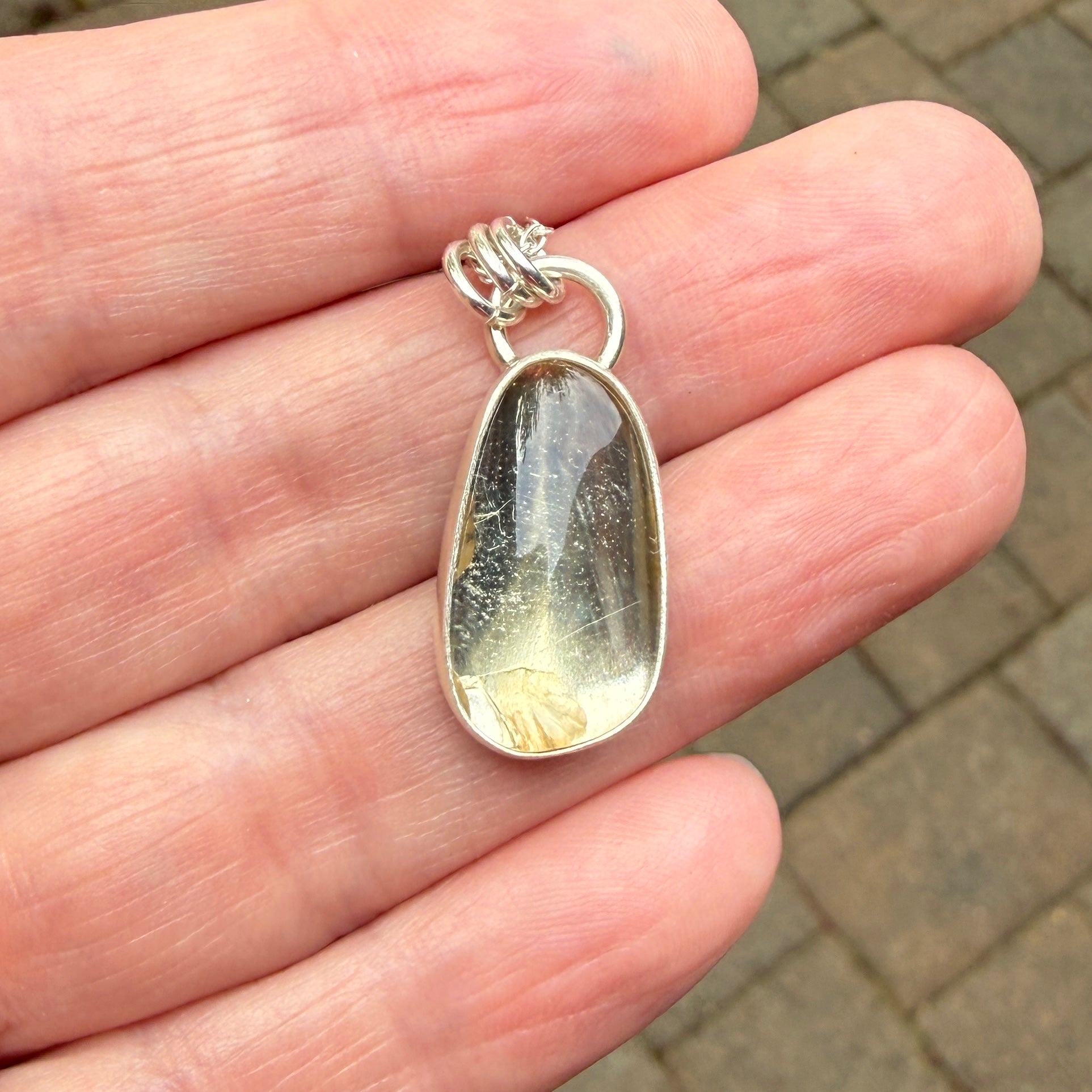 Silver pendant with a clear stone held in a hand on a blurred pavement background