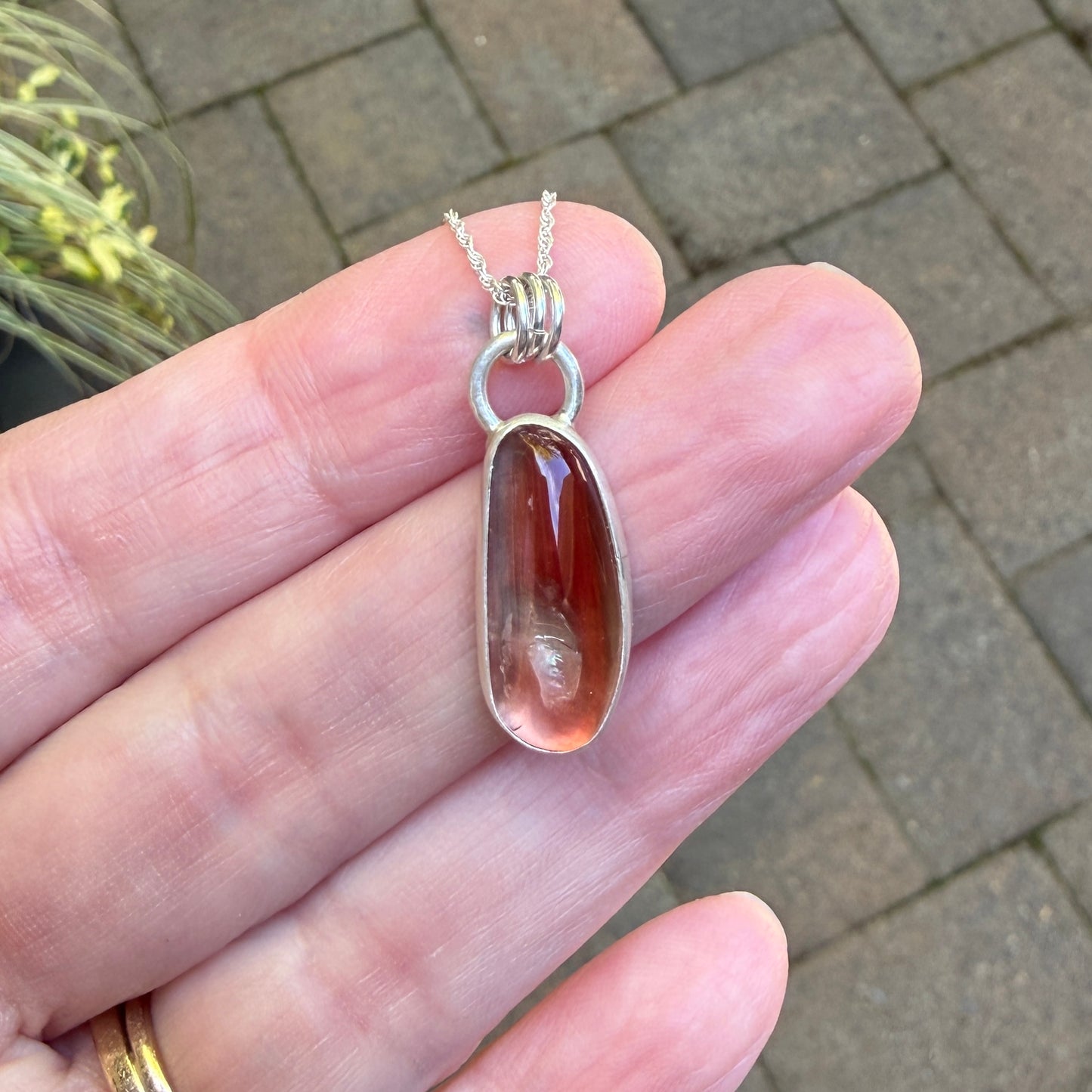 Hand holding a silver oregon sunstone pendant with a red stone against a pavement background