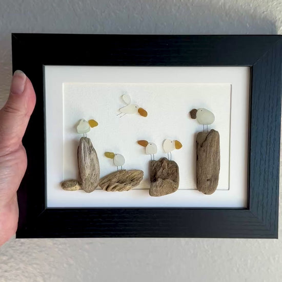 Framed sea glass artwork made of sea glass and driftwood on a white surface.