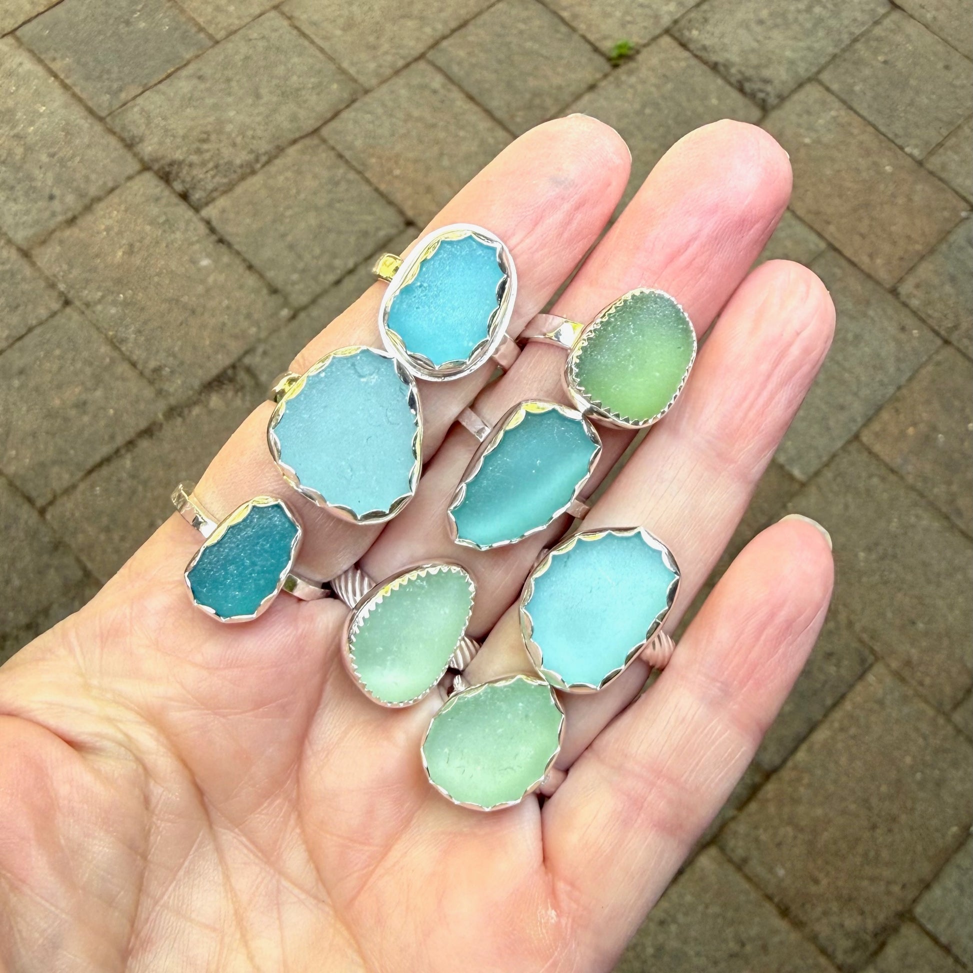 Hand holding multiple blue and green sea glass ring sets on a brick pavement background