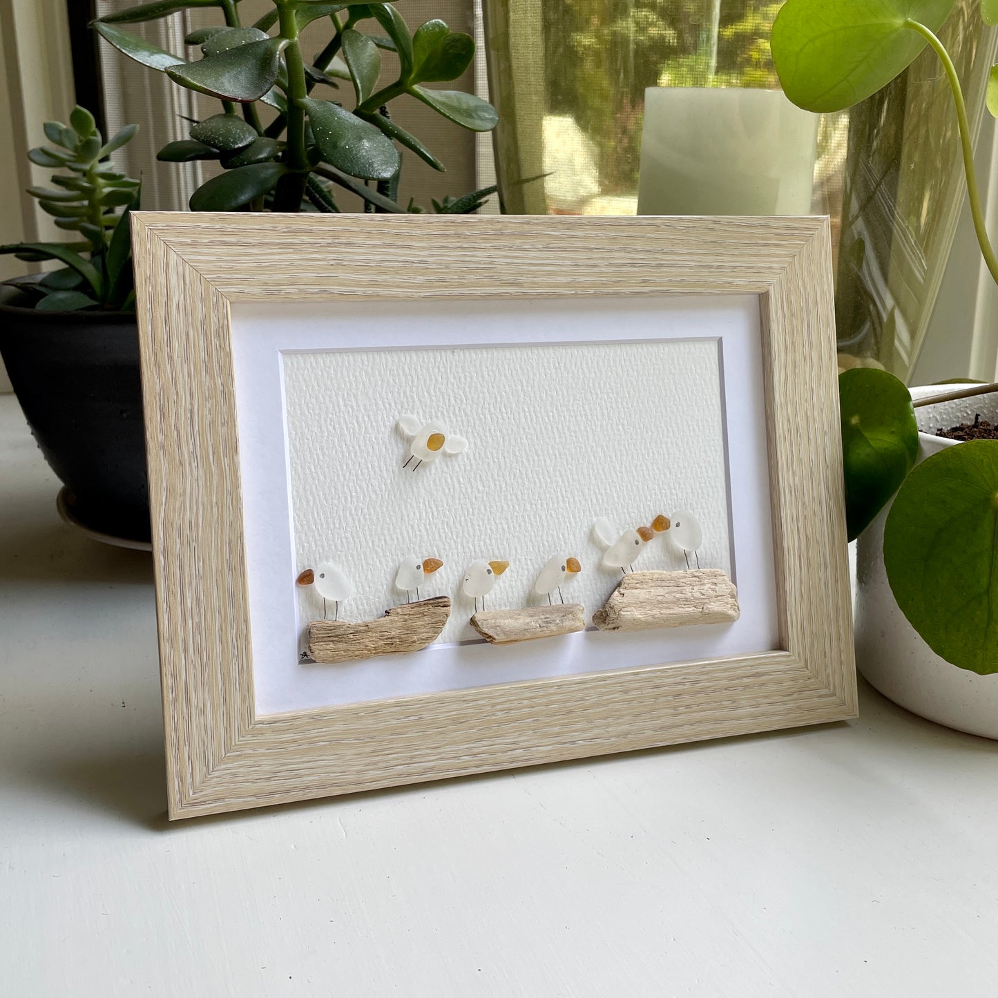 This is a framed 5 x 7 inch sea glass art picture featuring seagulls and driftwood logs. This item is perfect for gift giving and is ready to hang or place on a shelf. 