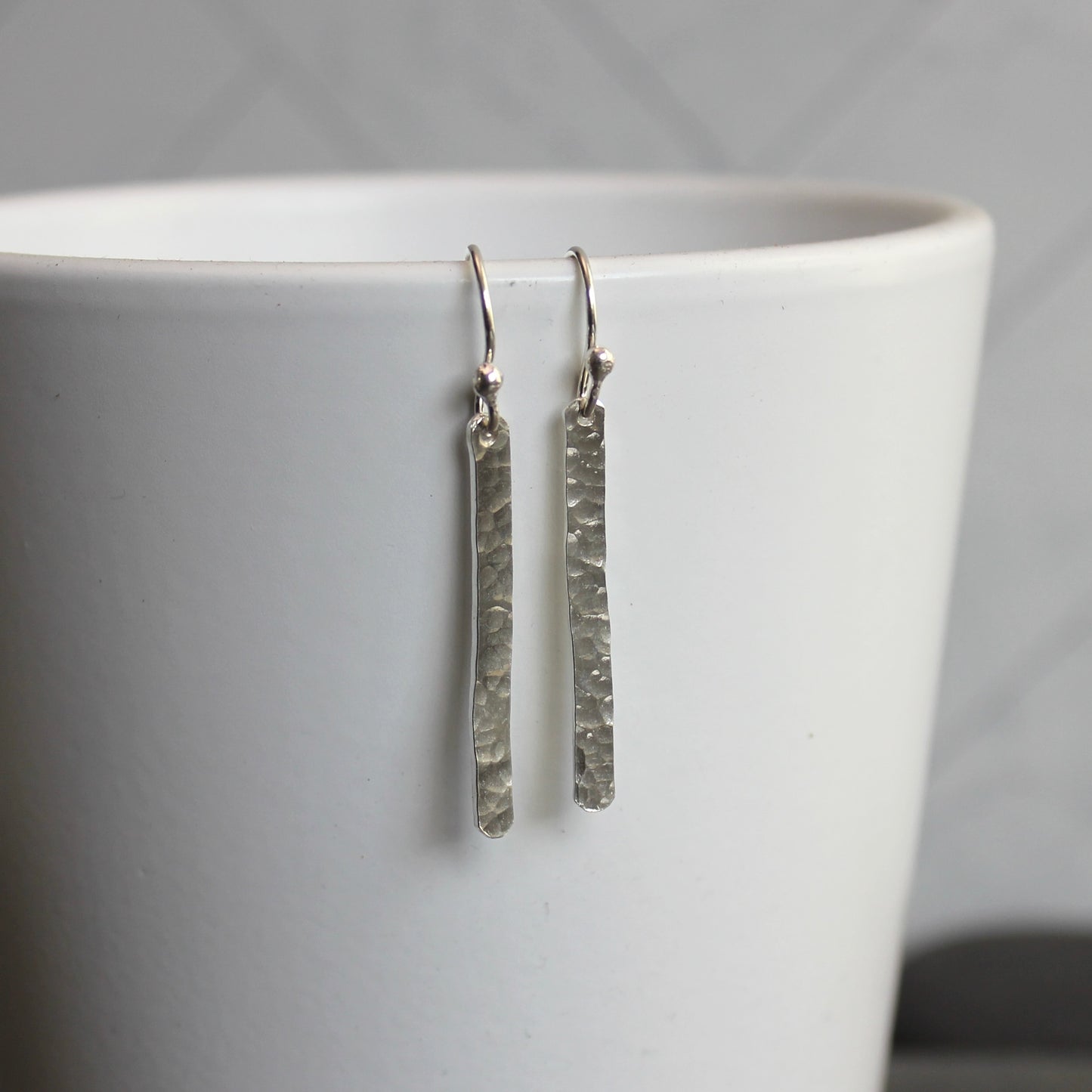 Silver textured earrings hanging from a white cup against a gray background
