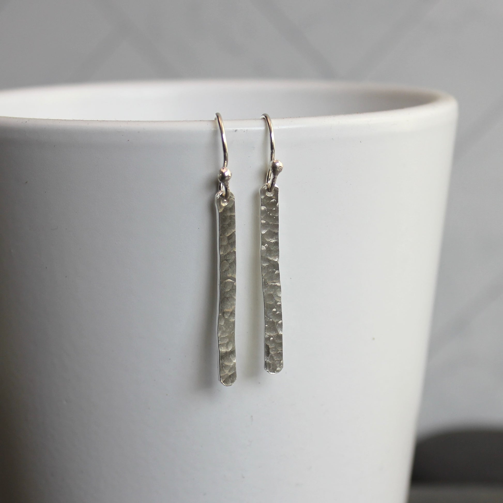 Silver textured earrings hanging from a white cup against a gray background