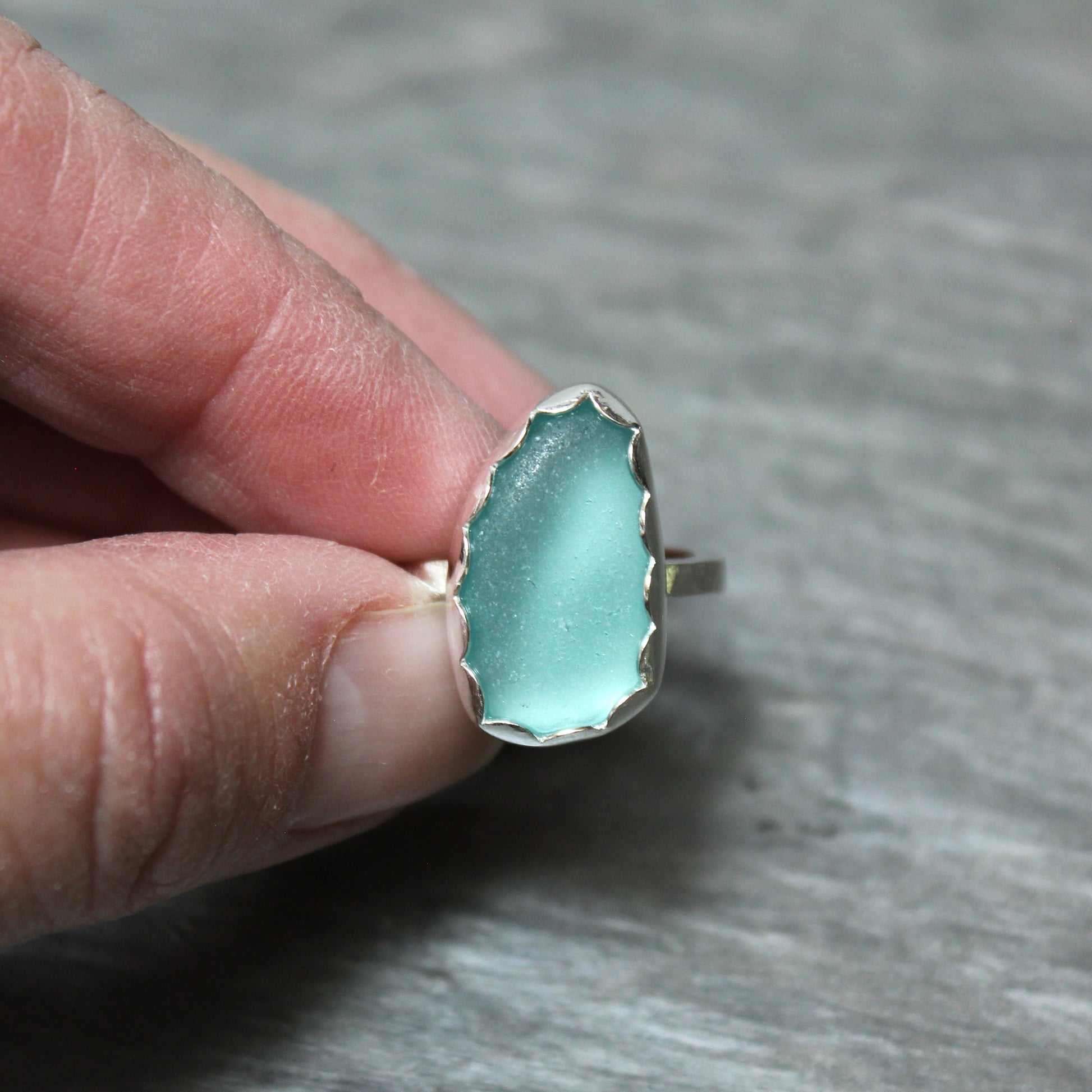 Turquoise sea glass ring with silver band held between fingers on a gray background