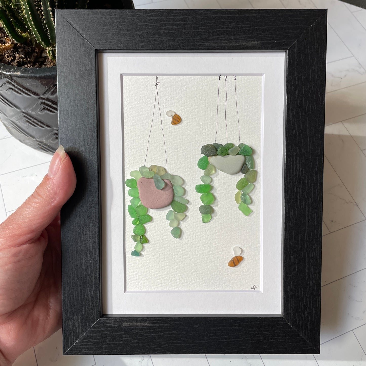 This 5 x 7 inch framed sea glass art picture features two hanging plants made from genuine sea glass, pottery & pebbles with a cute little sea glass bee buzzing about.  Designs may vary. 